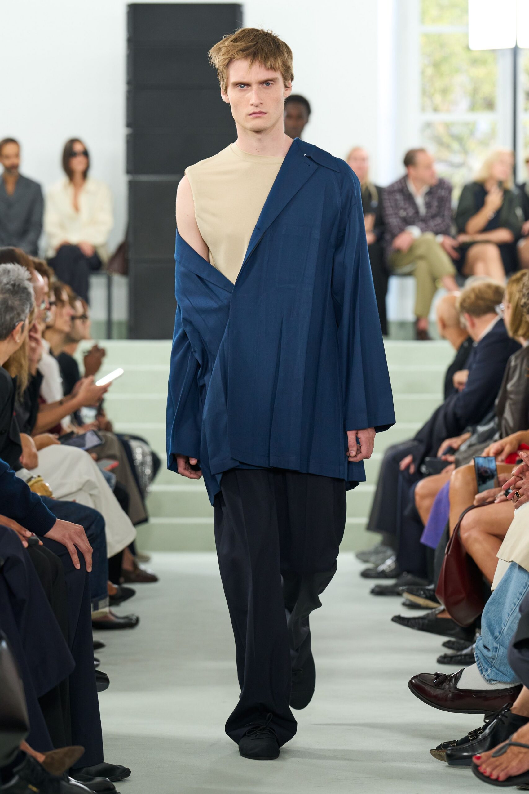Skin-close beige sleeveless top with a clean bonded-looking neck. Ink navy blazer worn half off, one shoulder dropped and twisted. Black fluid trousers breaking long over the shoe. Soft black slip-ons with rounded, almost slipper-like toes. Whatever you pull on for the hotel corridor ice machine at dawn.