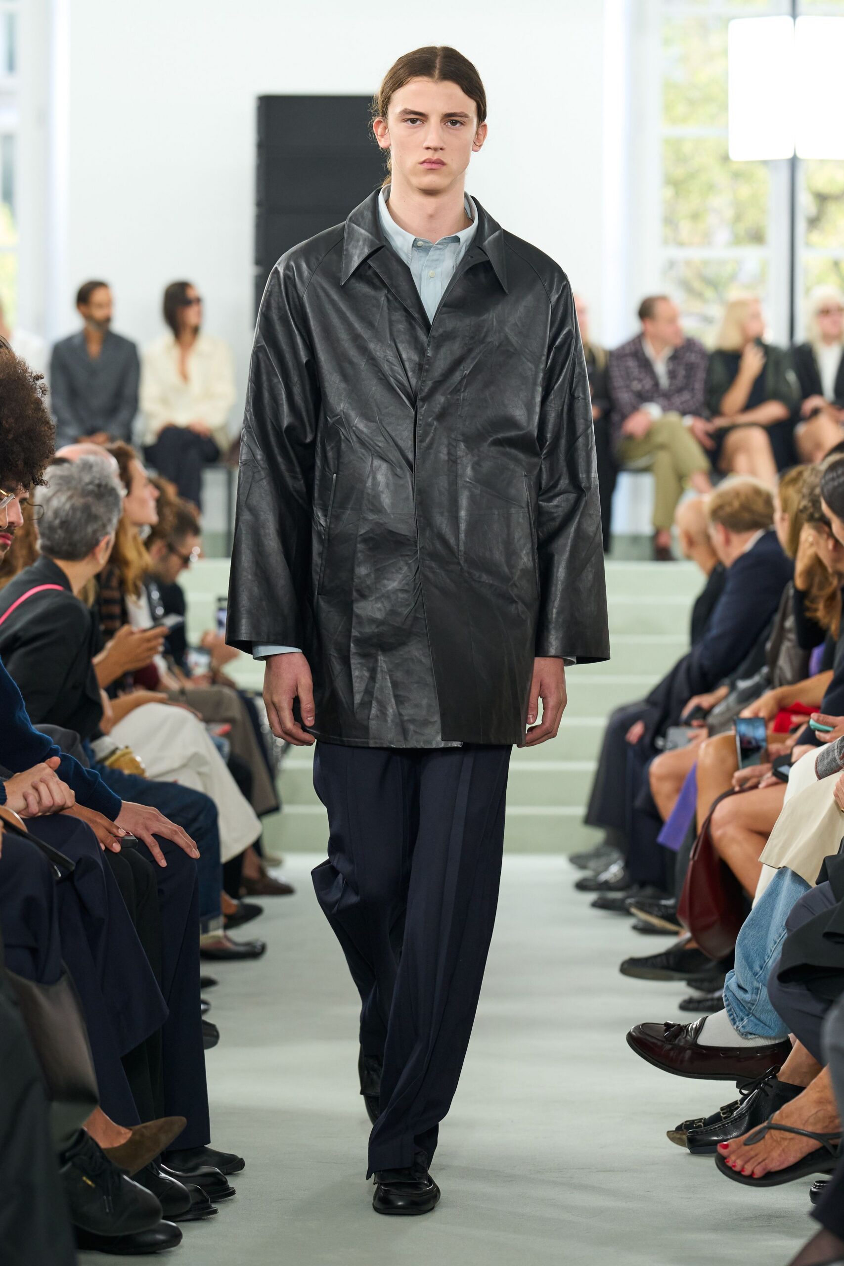Black leather coat, creased surface and dropped shoulders swallowing the frame. Pale blue shirt buttoned neat under the wide collar. Dark navy trousers falling in a long soft pleat. Black square-toe shoes. For crossing an echoing office lobby before anyone else has arrived.