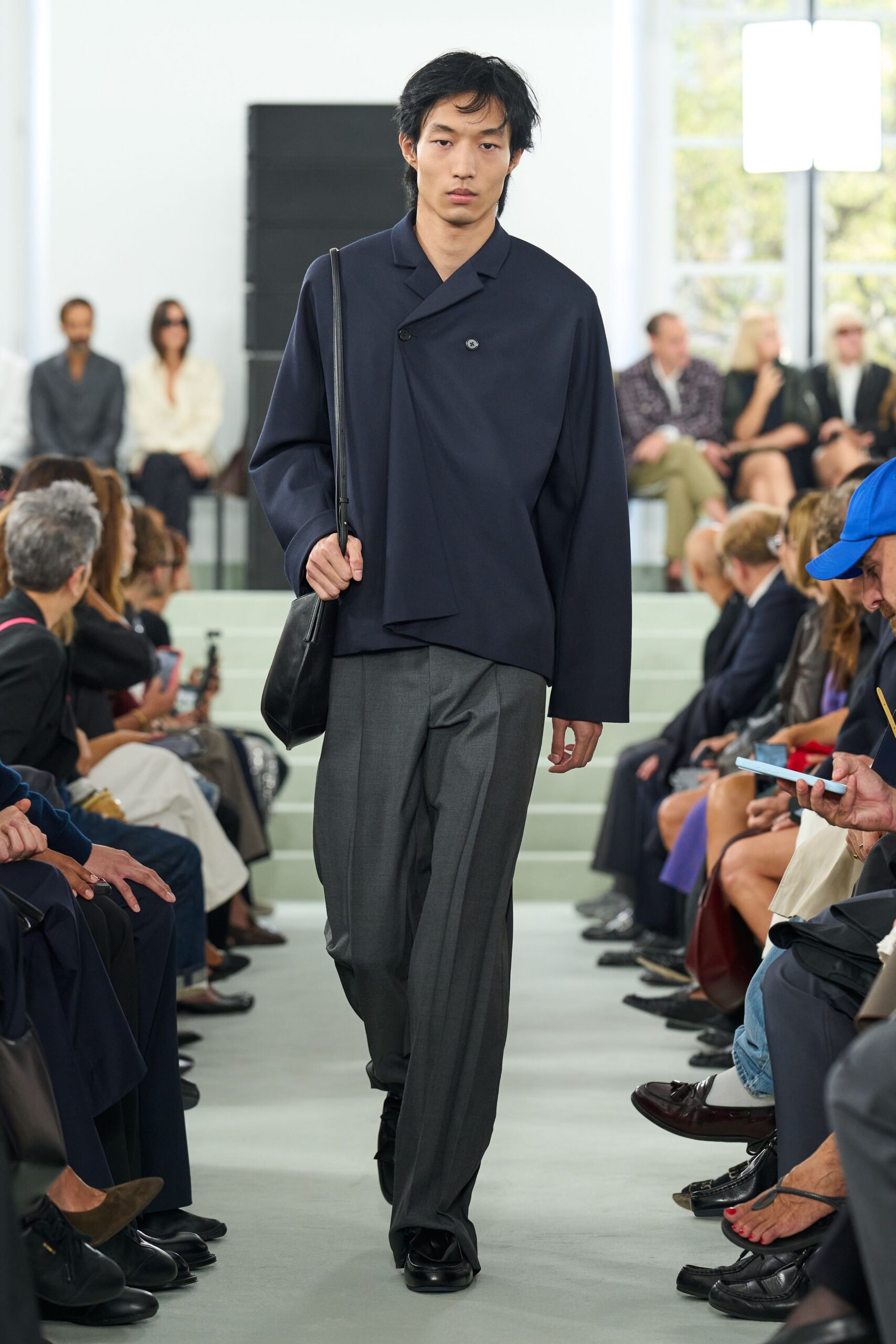 Ink navy wrap-front jacket with one off-center button and a falling panel. Black leather shoulder bag on a long narrow strap. Charcoal wool trousers with a pressed crease breaking over the shoe. Black square-toe shoes. Walking into a civic hearing with your notes folded in one pocket.