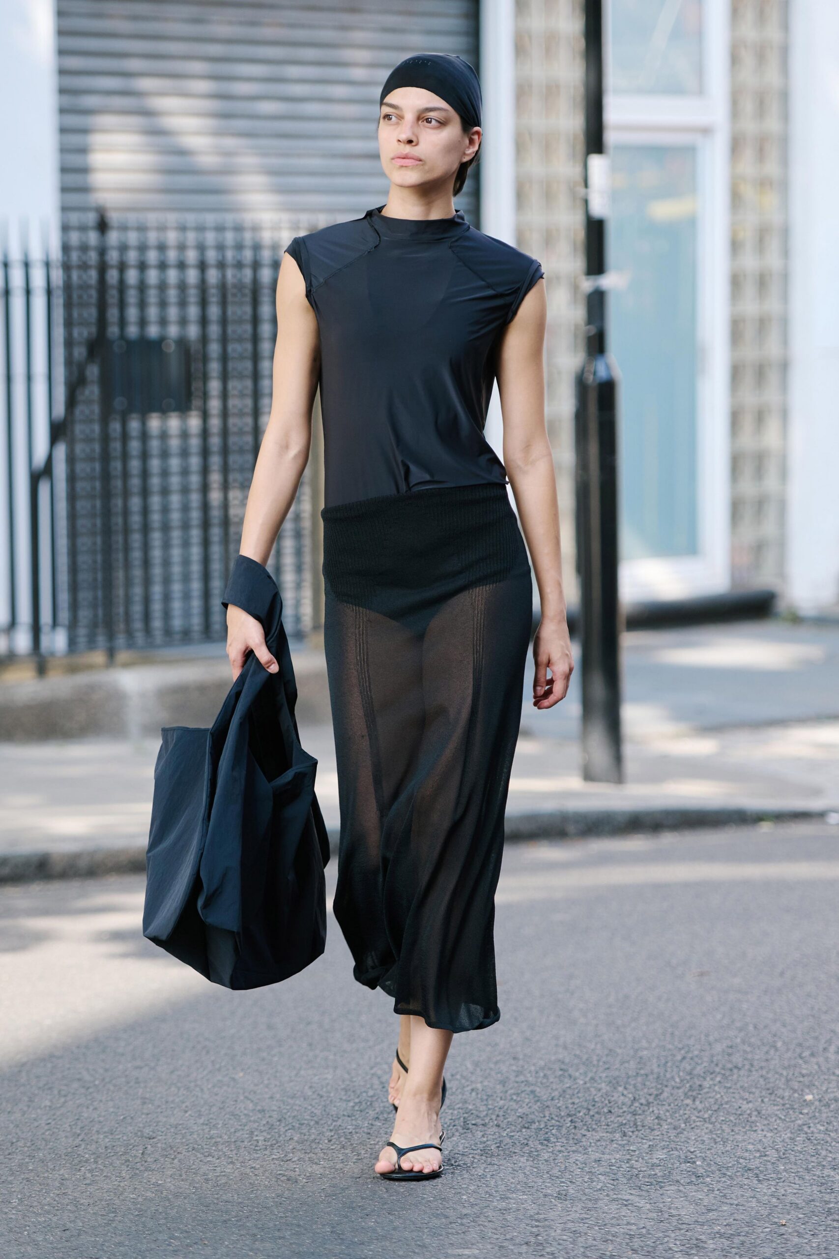 Black stretch headscarf pulled smooth over the crown. Inky jersey mock-neck top with cap sleeves and curved seams. Sheer black column skirt over a ribbed knit mini underlayer. Crumpled nylon tote hanging from one hand. Barely-there thong sandals with a slim heel. Crossing a quiet side street before the studios open.