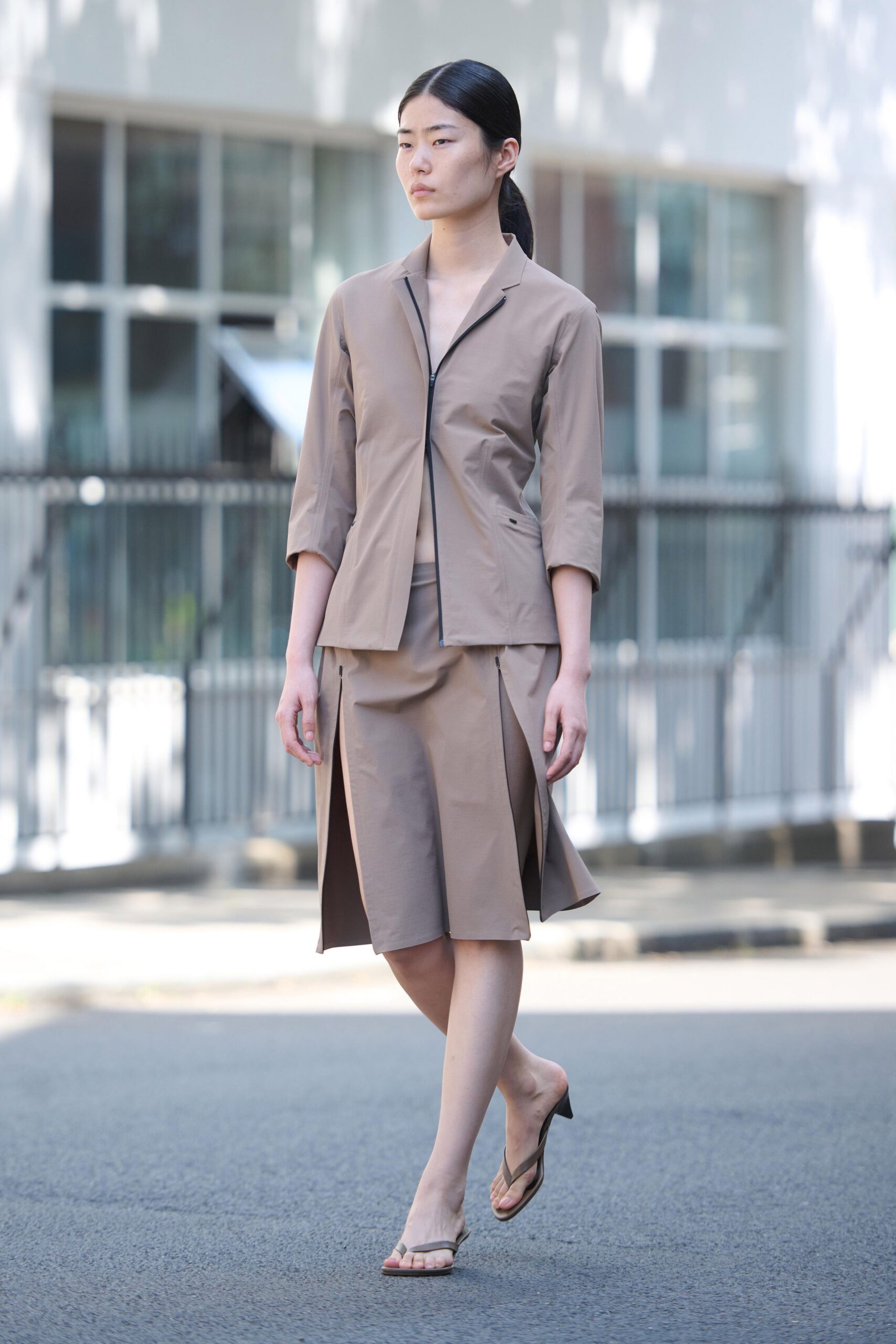 Taupe stretch jacket with an off-center black zip and bracelet sleeves. Matching midi skirt cut with deep front box pleats opening at the stride. Taupe thong sandals on a low kitten heel. Crossing the office plaza before the glass doors unlock.