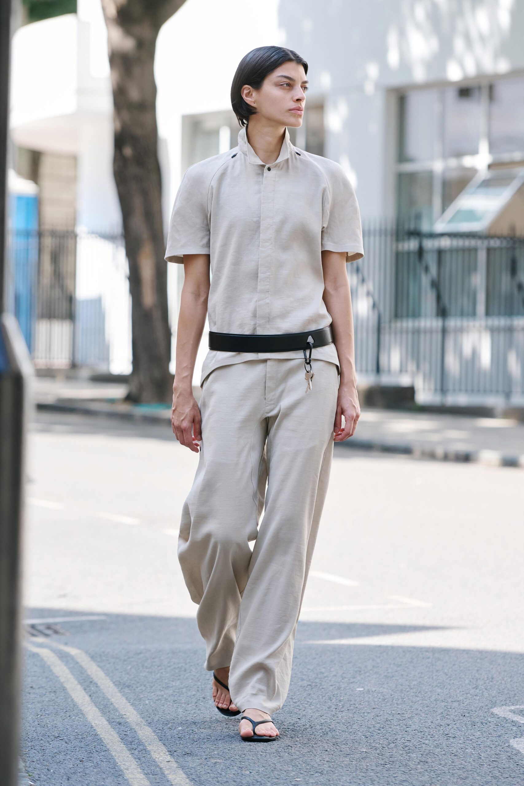 Pale stone short-sleeve top with a concealed placket and standing collar. Wide black belt with keys clipped to one side. Matching fluid trousers falling full and straight from the hip. Black thong sandals, almost flat. Crossing a quiet embassy street before the heat settles in.