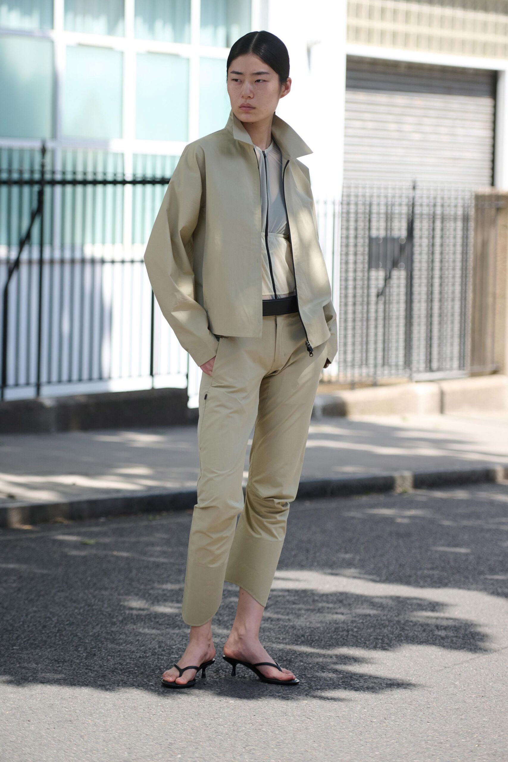 Pale khaki shell jacket, stand collar and black zip tracing off-center. Sheer cream mesh top under a fitted zip-front panel. Matching tapered shell trousers, a tiny zip pocket set on the thigh. Black thong mules with narrow kitten heels. Crossing the side street after lunch, office badge still in your pocket.