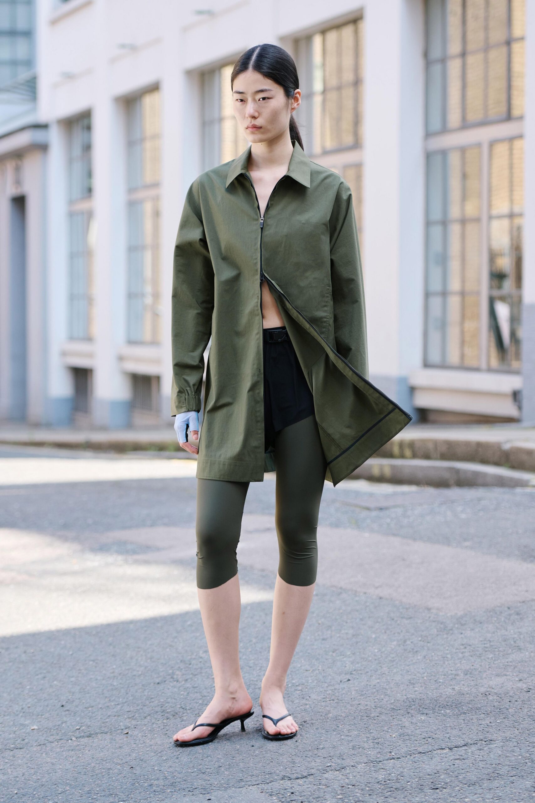 Olive cotton zip-front shirt, one side kicked open from the hem. Black shorts peeking from beneath the split placket. Olive stretch leggings cropped below the knee. Pale grey fingerless glove on one hand. Black thong mules with a low kitten heel. Walking across the service road before the studios unlock.