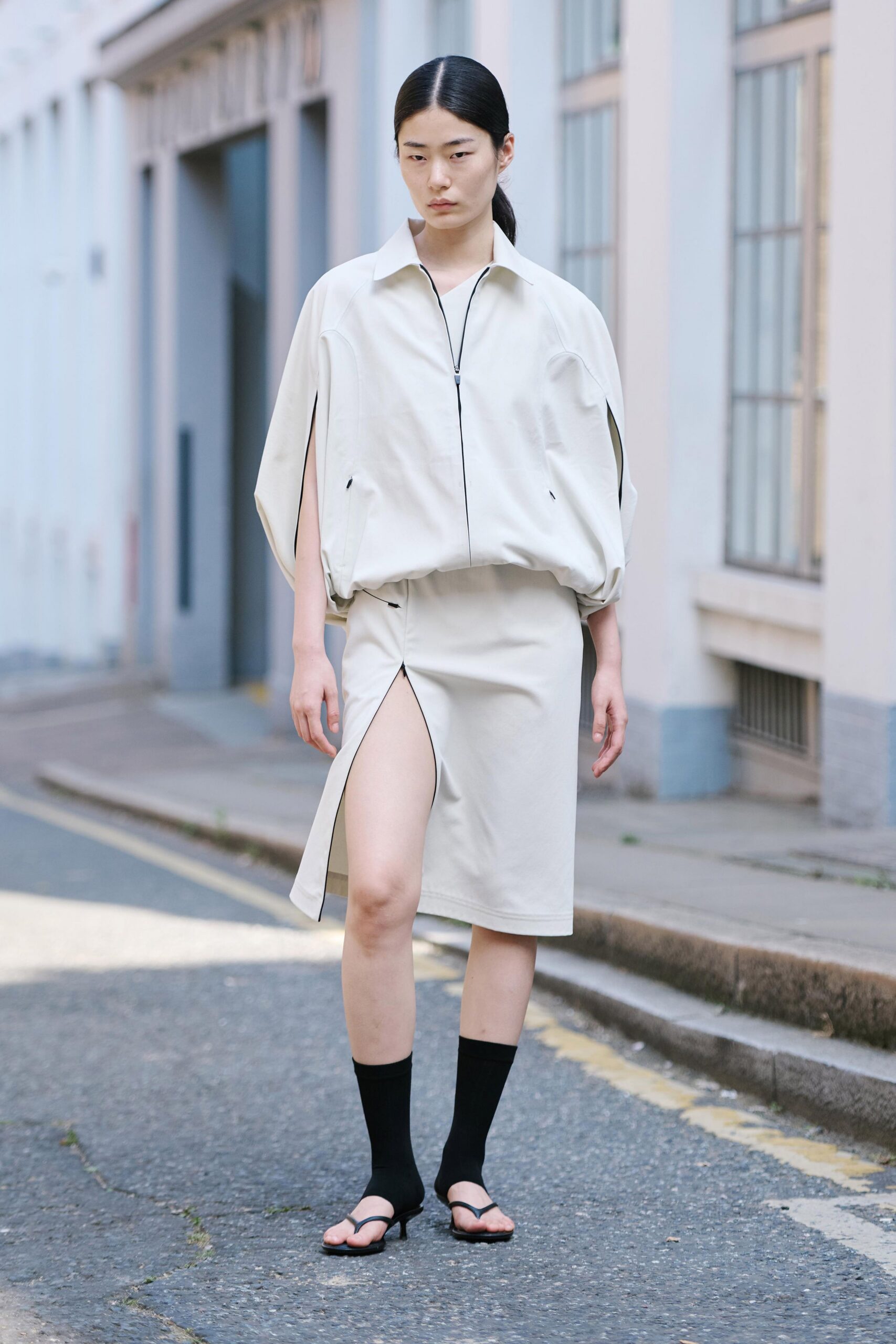 Pale stone zip-front top, ballooned hem and slit cape sleeves. Matching column skirt with a high front slit edged in black piping. Black ankle socks. Black thong kitten heels. Crossing the side street outside a clinic before the heat settles in.