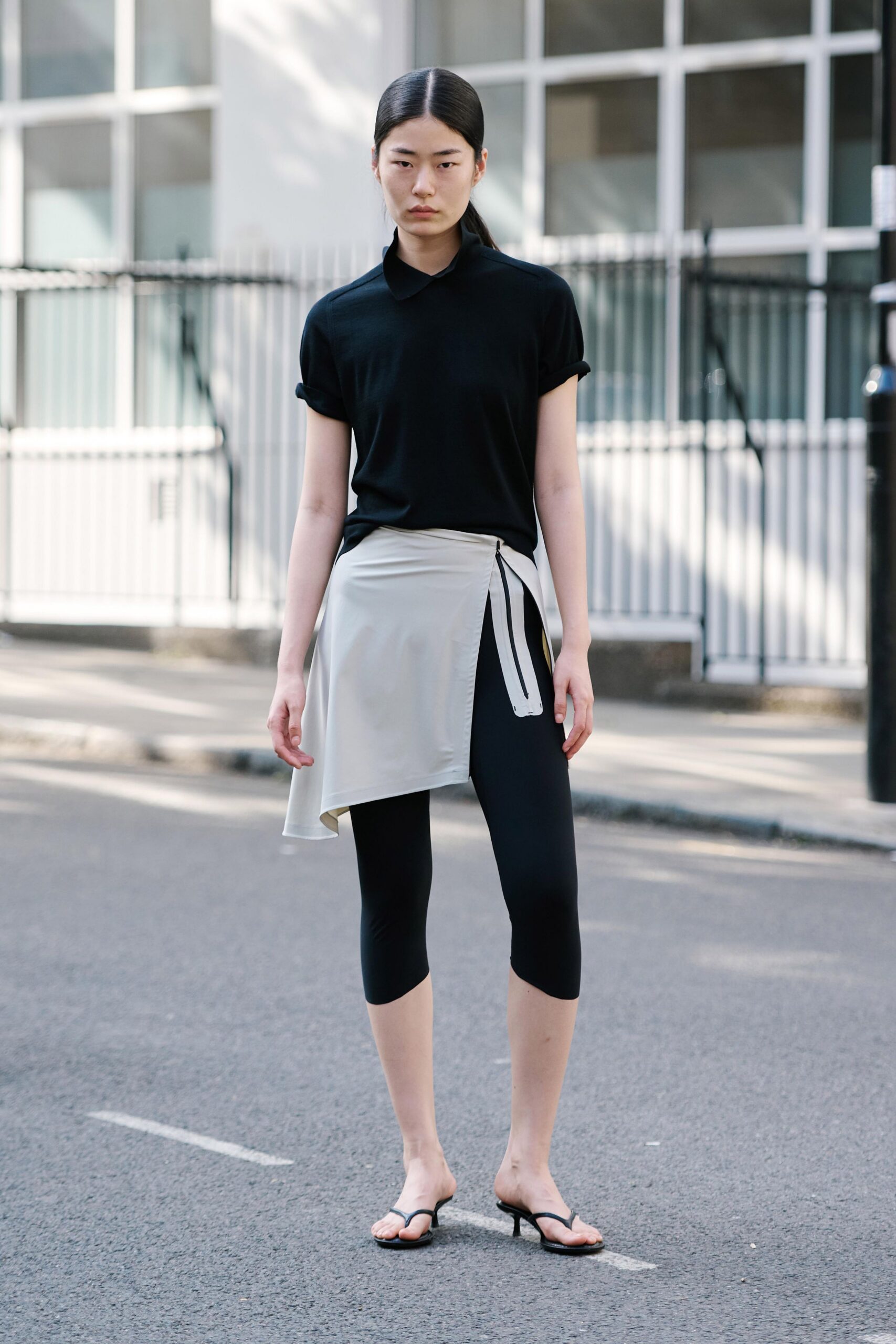 Black fine-knit polo, sleeves rolled into neat cuffed tubes. Chalk wrap skirt panel hanging unevenly over the hip. Black cropped leggings, second-skin stretch to mid-calf. Low black thong mules with a narrow kitten heel. Crossing a side street before the office towers throw full shade.