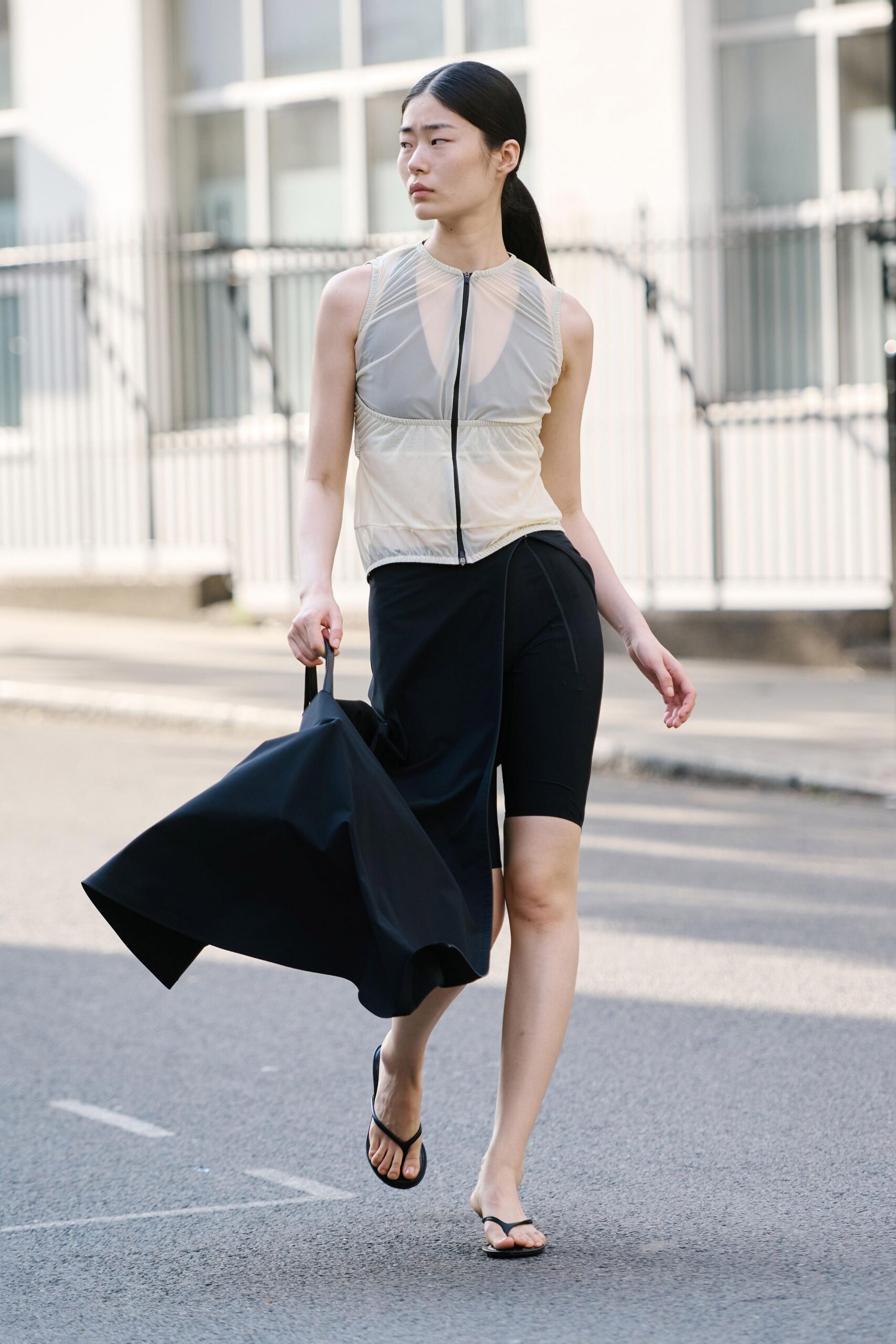 Sheer oyster mesh top with a black zip splitting the ruched front. Black stretch shorts, one leg left fully exposed. Asymmetric black overskirt falling in a stiff curved panel. Huge black tote collapsing into sharp origami folds. Flat black thong sandals. Crossing a quiet embassy block with dry-cleaning and nowhere urgent to be.