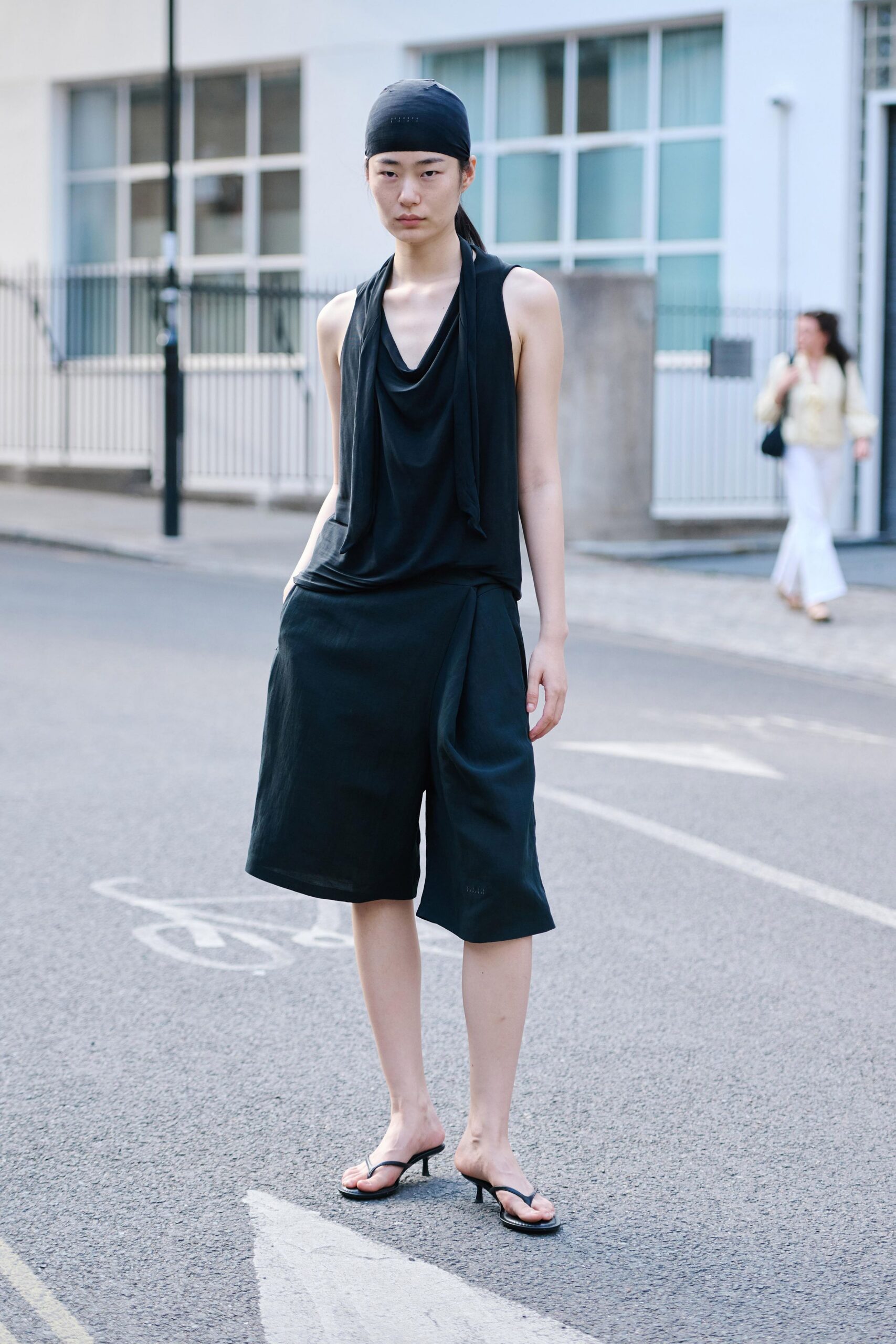 Black stretch headscarf pulled smooth, tiny perforations near the crown. Black jersey draped in a deep cowl, long scarf panels falling straight. Matte black culottes with a wrapped front fold breaking at one knee. Barely-there thong mules on short pin heels. Crossing a quiet side street before the galleries open.