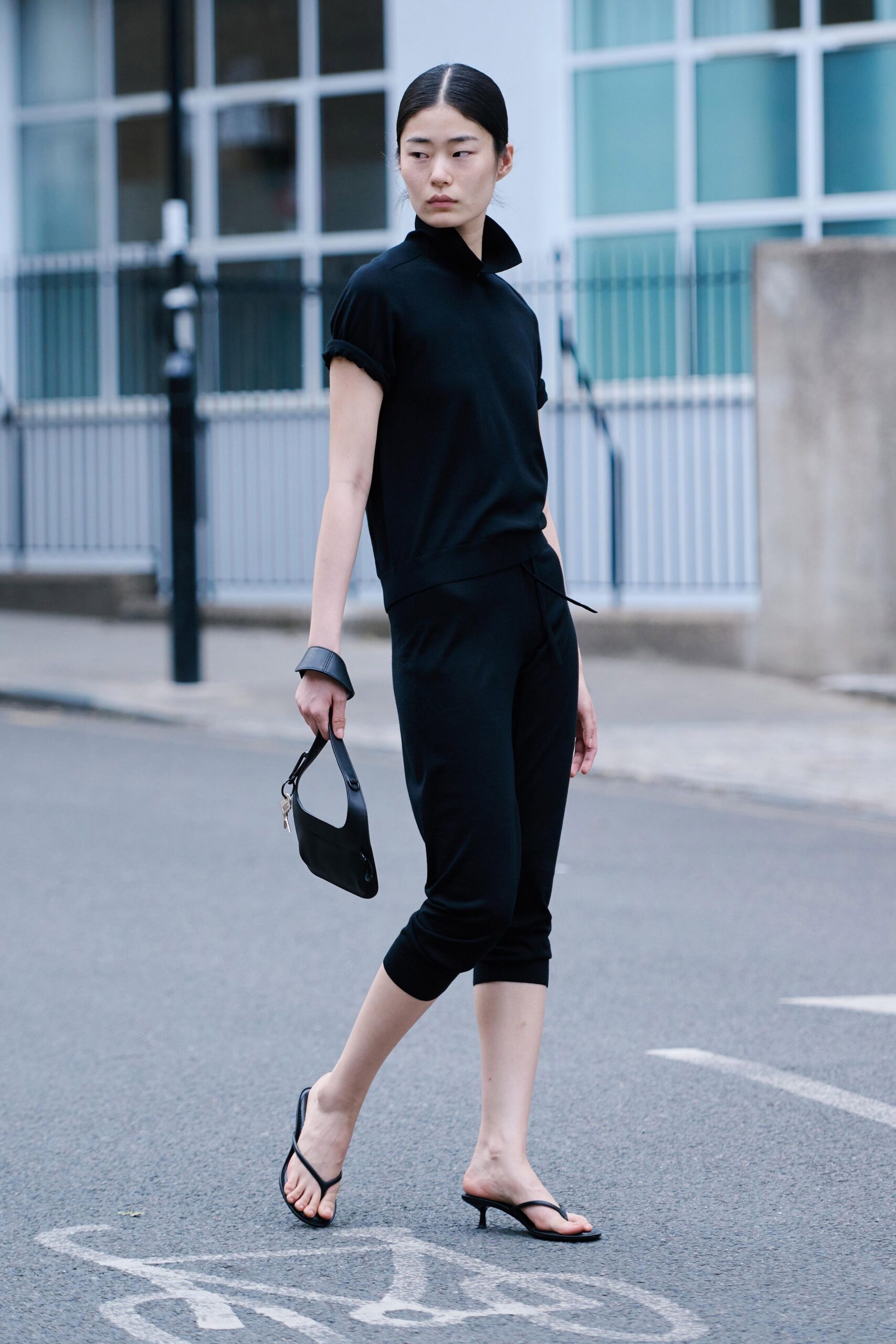 Black knit top with a high stand collar and rolled cap sleeves. Wide black cuff, smooth and slightly curved. Matching knit capris, tapered to a close calf cuff. Small black shoulder bag with keys clipped at the handle. Black thong mules on a low kitten heel. Crossing a side street before the office crowd spills out for lunch.