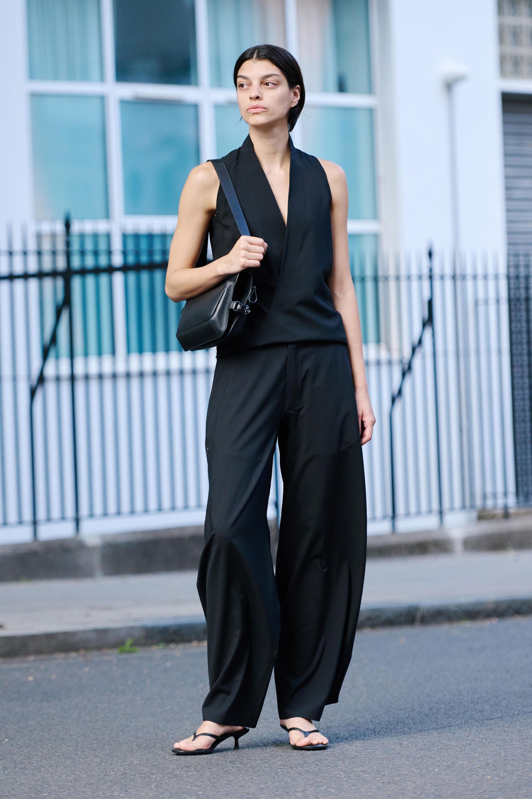Black sleeveless wrap top with a deep shawl-collar plunge. Structured black leather bag with a clipped side strap. Fluid black wide-leg trousers, pressed flat then breaking at the ankle. Black thong kitten heels. Crossing a quiet embassy street before the office air-conditioning kicks in.