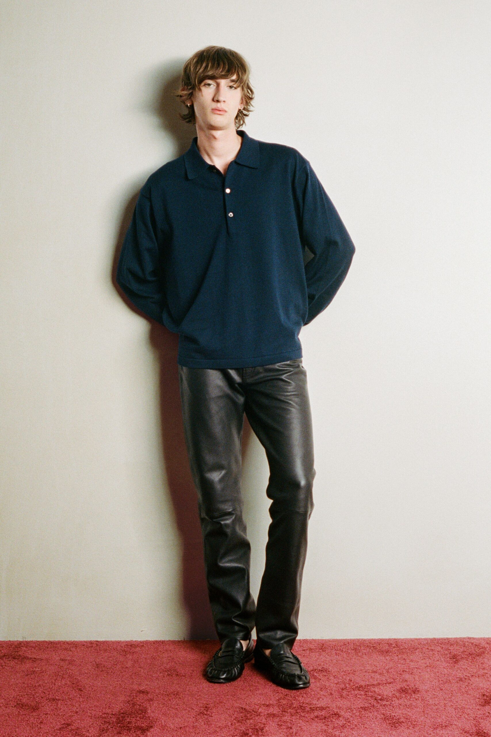 Ink-navy knit polo with a loose placket and two pale buttons. Black leather trousers cut narrow, creasing softly at the knee. Black loafers with gathered apron seams and a low vamp. Hotel hallway carpet after midnight, ice bucket in someone else's room.