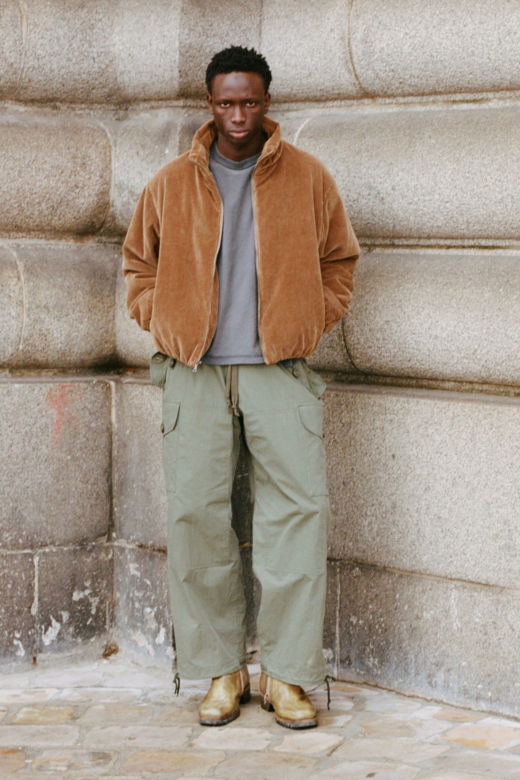 Camel velour zip jacket, collar standing high and hem softly bloused. Washed grey crewneck tee peeking flat beneath the open front. Sage cotton cargos cut wide, flap pockets and dangling hem ties. Bronzed leather ankle boots with a squared toe and dark welt. Cold pavement outside a stone post office just before it opens.