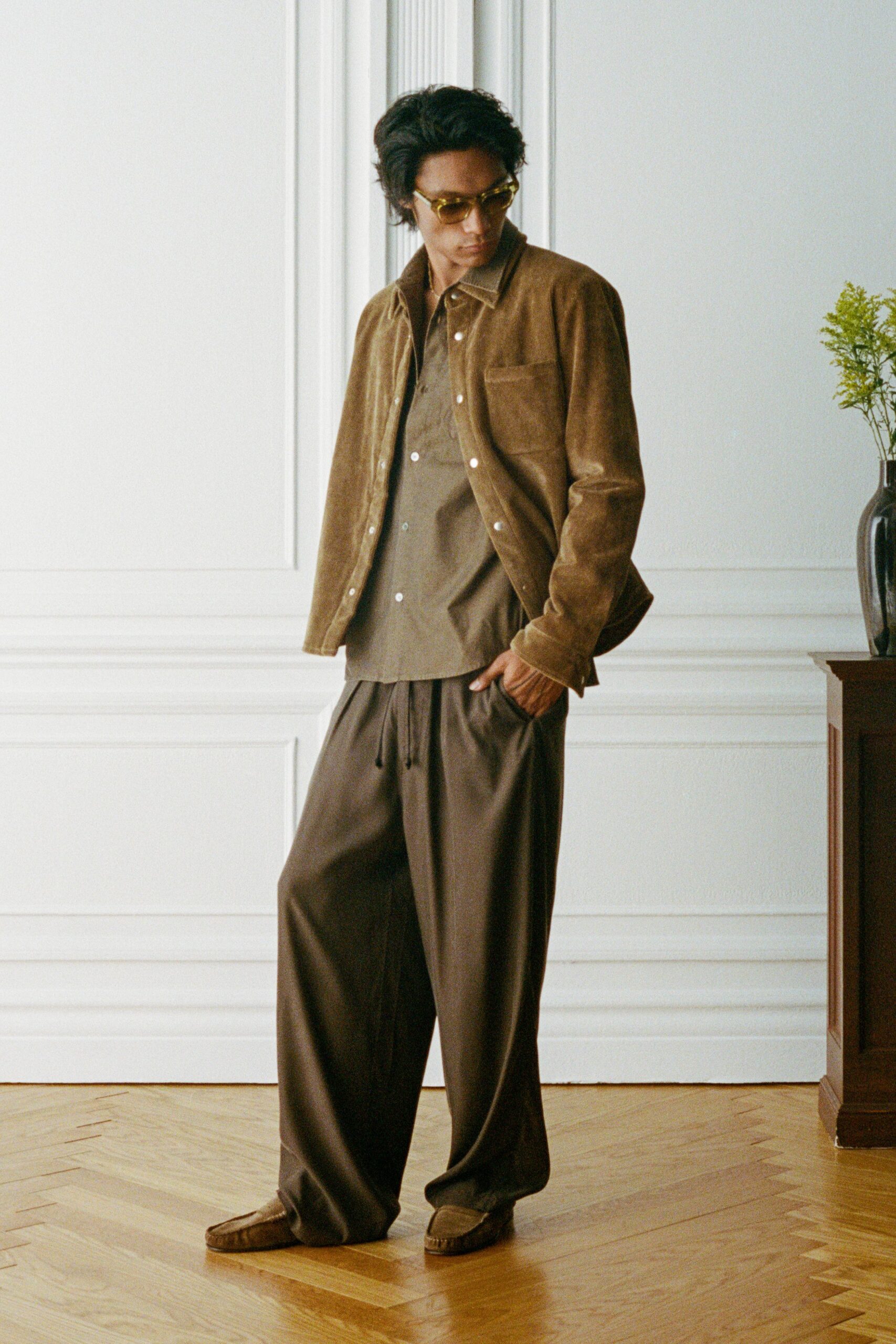 Honey-tinted acetate frames. Tobacco suede shirt jacket with bright pearl snaps. Moss button-front shirt, soft point collar sitting open. Dark draped drawstring trousers, pooling over the vamp. Brown suede loafers with raised apron stitching. Afternoon in a rented salon, flowers dropped off too early.
