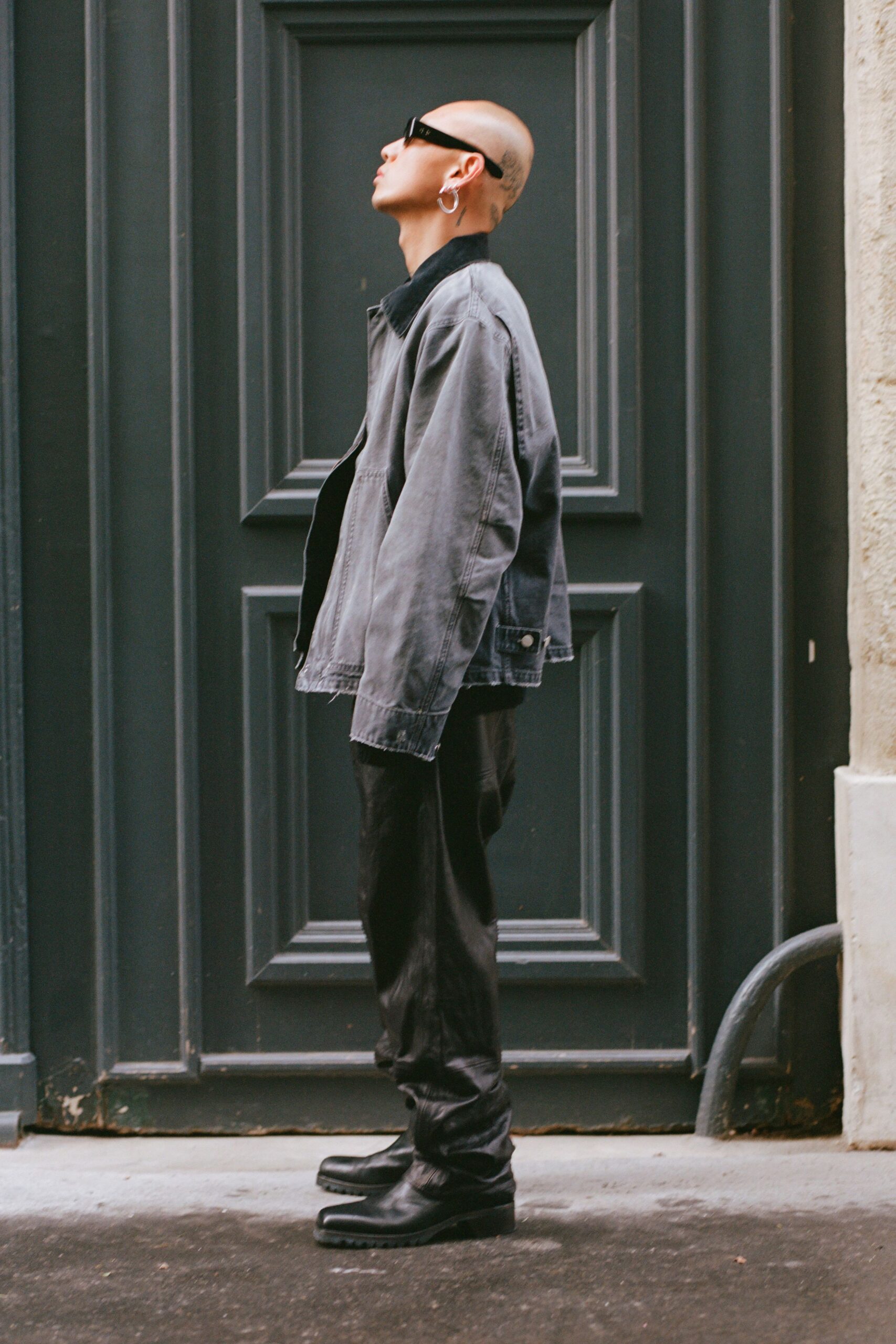 Narrow black wraparound sunglasses. Mismatched silver hoops at one ear. Washed charcoal denim jacket with a black corduroy collar and frayed hem. Black trousers falling long and pooling over the instep. Square-toe leather boots with deep lug soles. Waiting outside a shuttered bar before the delivery truck arrives.
