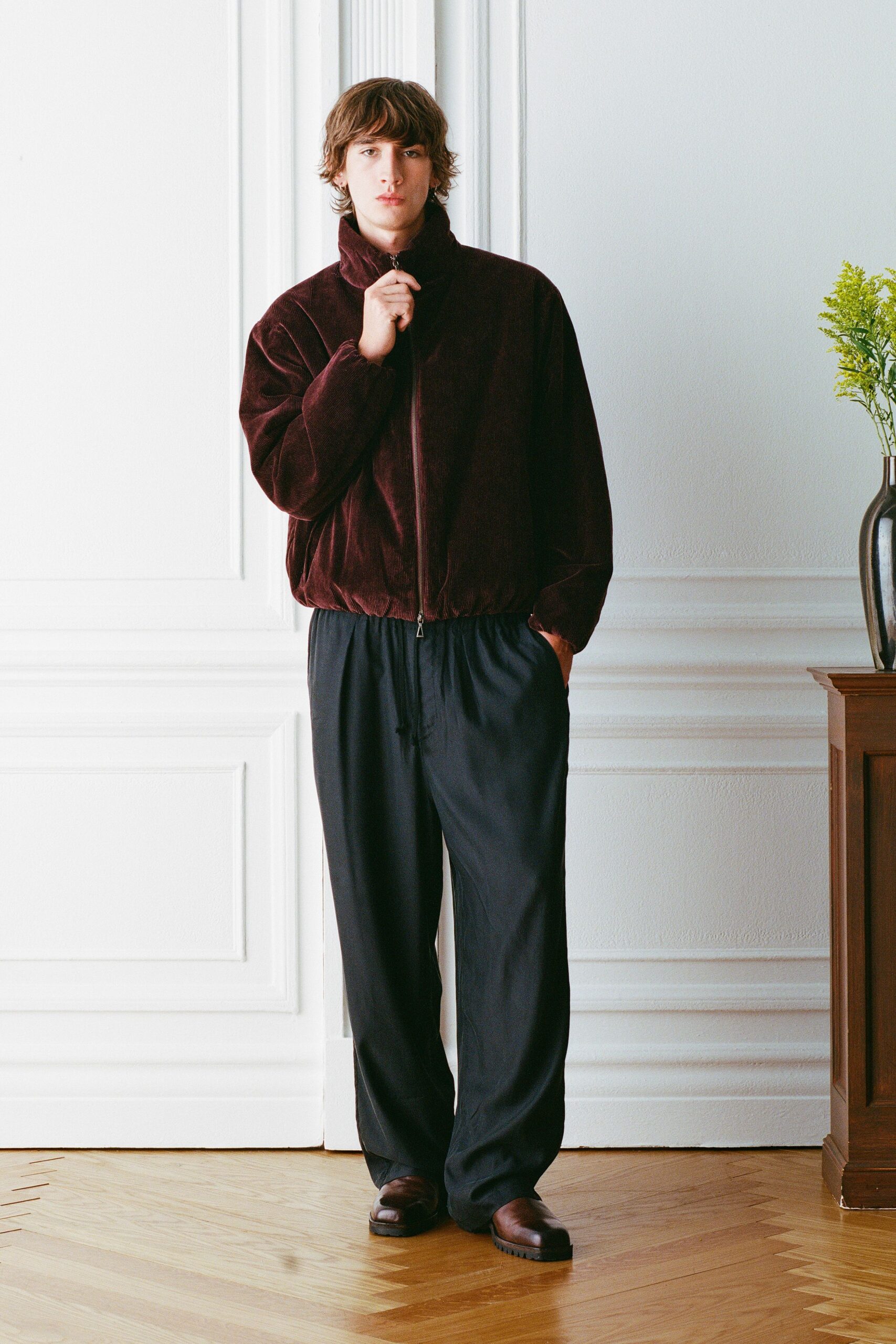 Burgundy corduroy blouson, stand collar zipped to the mouth. Black drawstring trousers in a fluid weave, cords hanging loose. Brown leather clogs with thick ridged soles. Sunday morning in a rented apartment, flowers already dropping pollen.