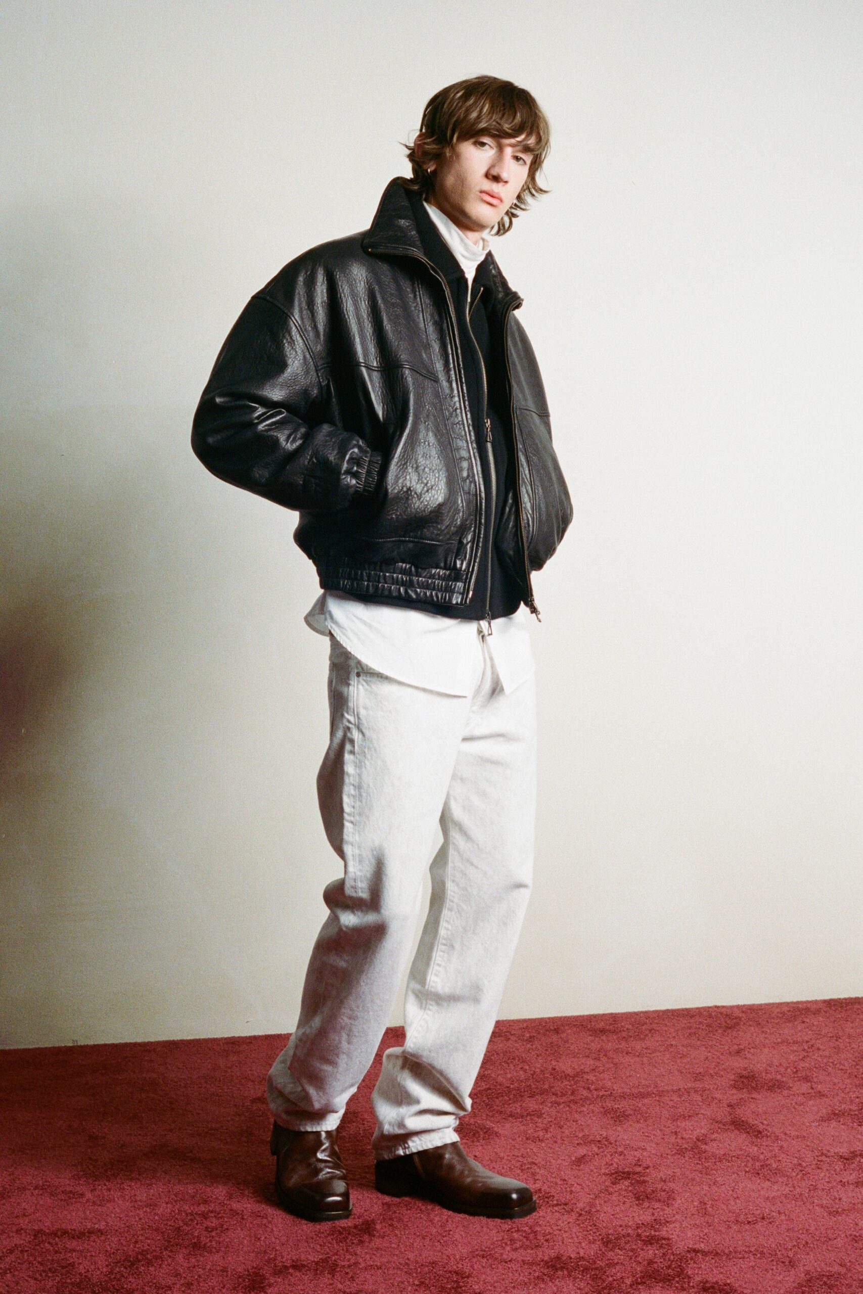 Black pebbled leather bomber, collar tipped upright above a two-way zip. White shirt hem hanging loose below the jacket. Chalk-wash denim cut straight and pooling lightly at the ankle. Dark square-toe leather boots with a blunt stacked heel. For crossing a hotel lobby before sunrise, keycard still warm in hand.