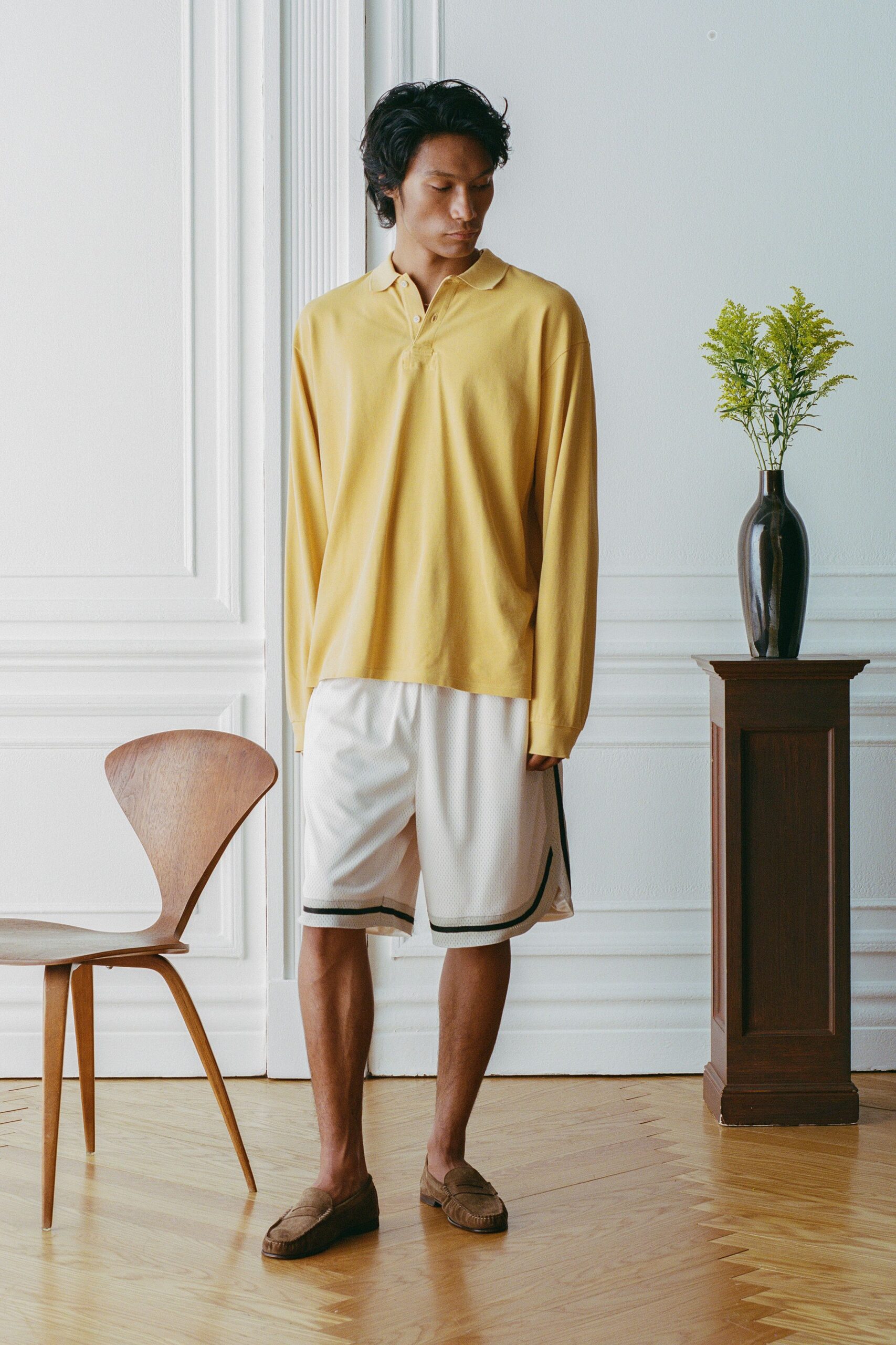 Butter-yellow jersey polo, dropped shoulders and a two-button placket left open. White mesh basketball shorts with black binding skimming the side slit. Brown suede penny loafers, softly creased across the vamp. Sunday in a rented apartment, florist stems still waiting for water.
