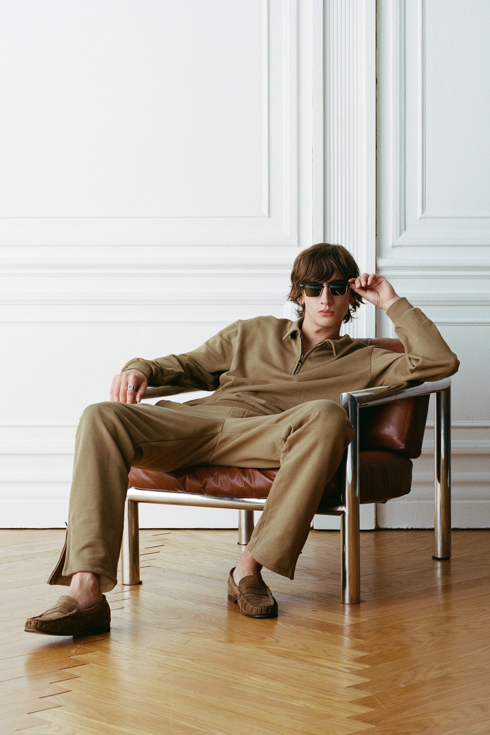 Black square sunglasses with thick temples. Taupe knit zip polo with a long pointed collar. Matching taupe trousers, pressed crease softening at the knee. Brown suede penny loafers with raised moc stitching. Chunky silver ring with a pale stone face. Afternoon in a borrowed apartment, waiting for the espresso to drip.