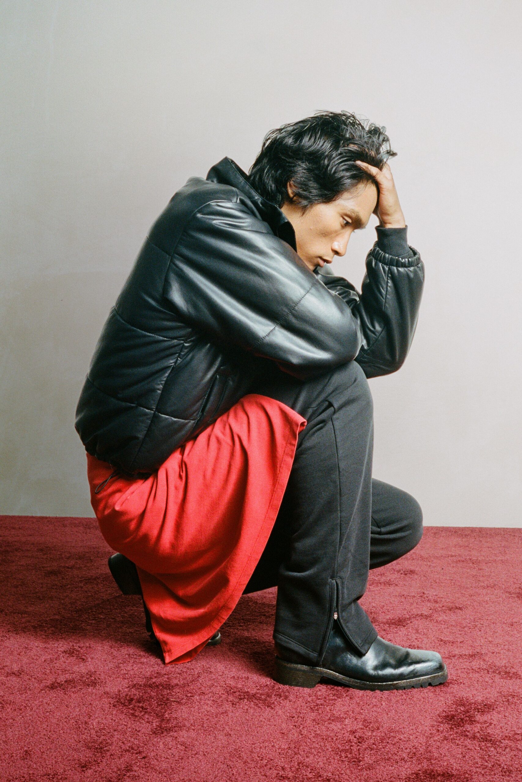 Black leather bomber, paneled and padded with ribbed knit cuffs. Tomato-red woven wrap falling in heavy folds over the knee. Black tailored trousers with a short ankle zip left open. Square-toe black leather boots with a thick stacked sole. Kneeling on the motel carpet after missing the last bus out.