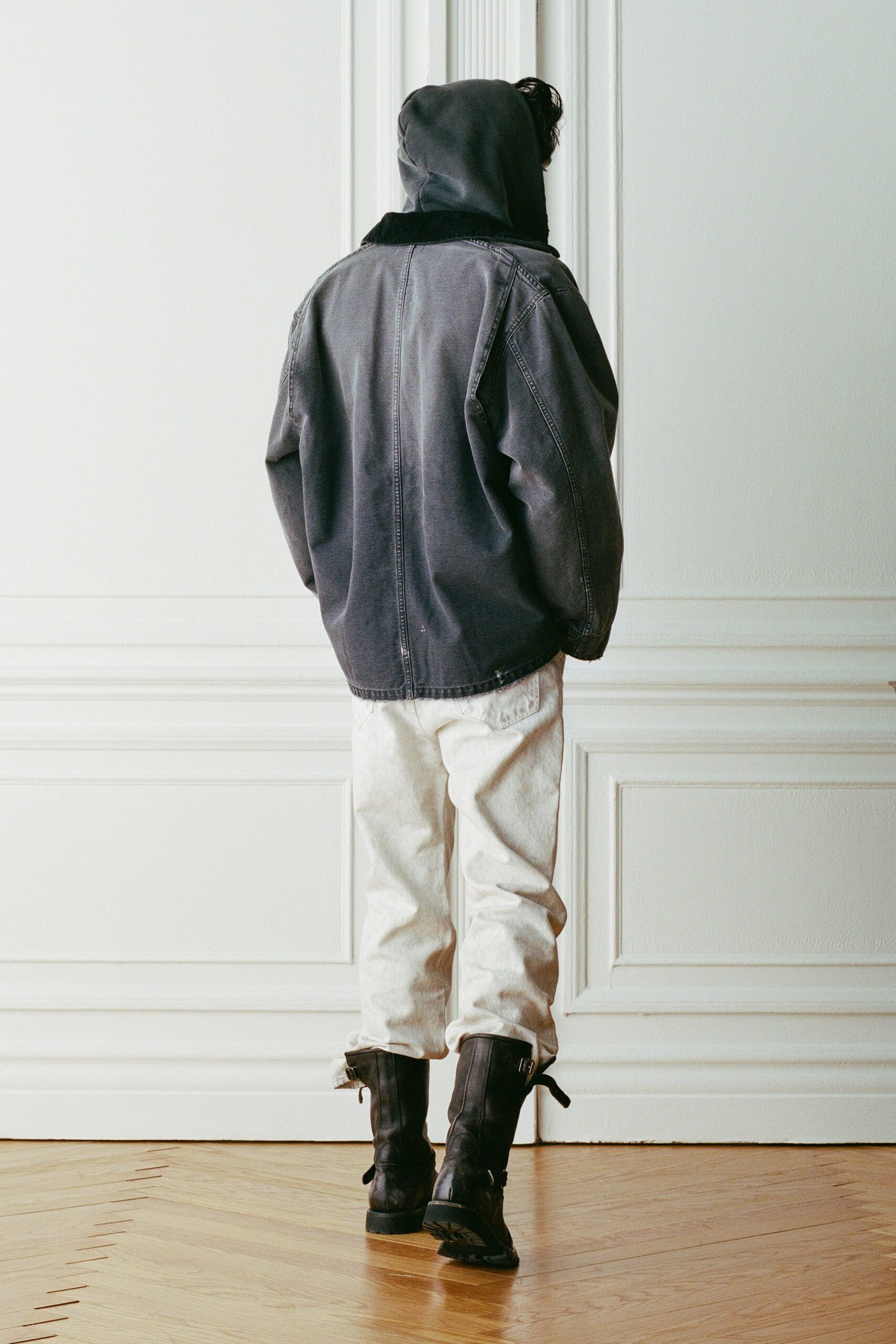 Charcoal hood pulled up, dense fleece standing thick at the neck. Washed black denim jacket, dropped shoulders and a scuffed hem. Chalk denim jeans, loose through the leg with a low back pocket. Black leather engineer boots with loose ankle straps. Crossing an empty apartment with the windows cracked after rain.