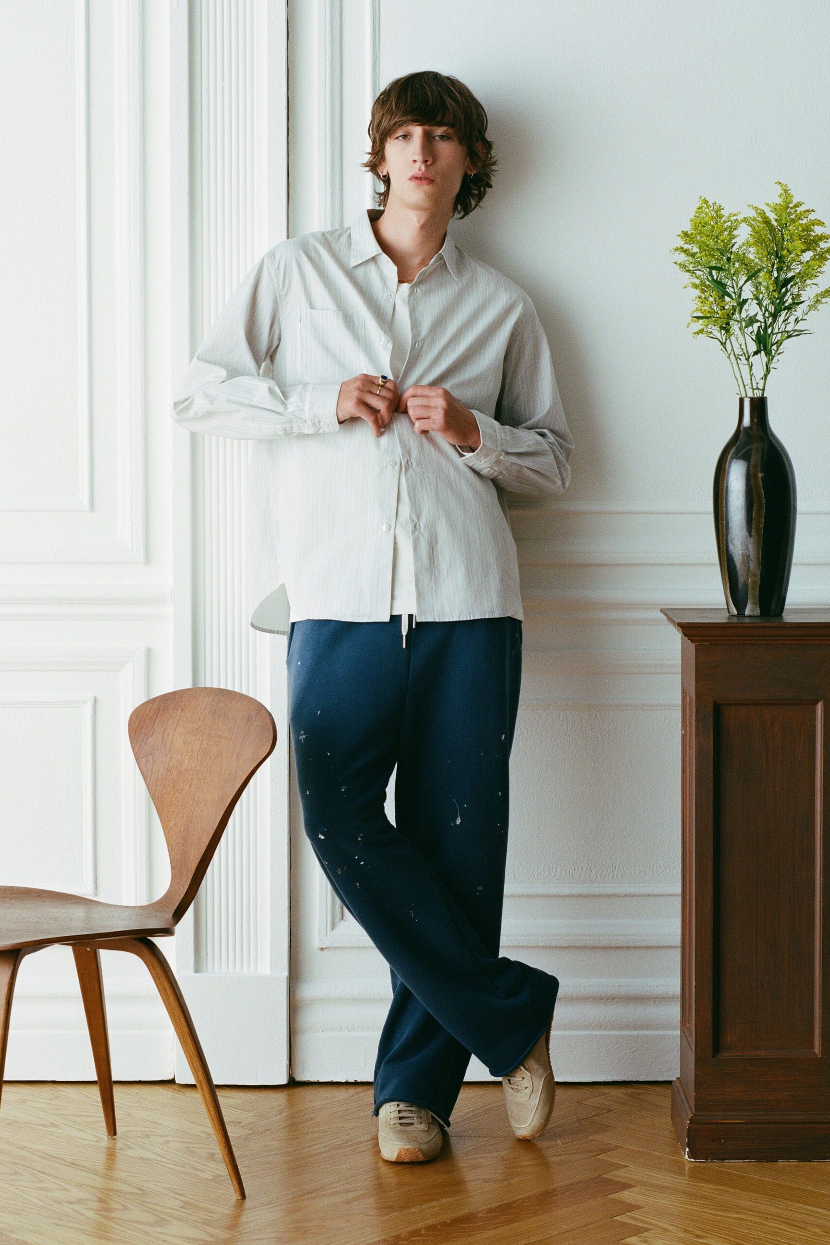 Pale striped cotton shirt with a soft crease and patch pocket. White crewneck tee peeking through the open placket. Navy jersey track pants speckled with white paint flecks. Off-white suede sneakers with gum peeking at the sole. Sunday morning in a rented apartment, flowers already on the sideboard.