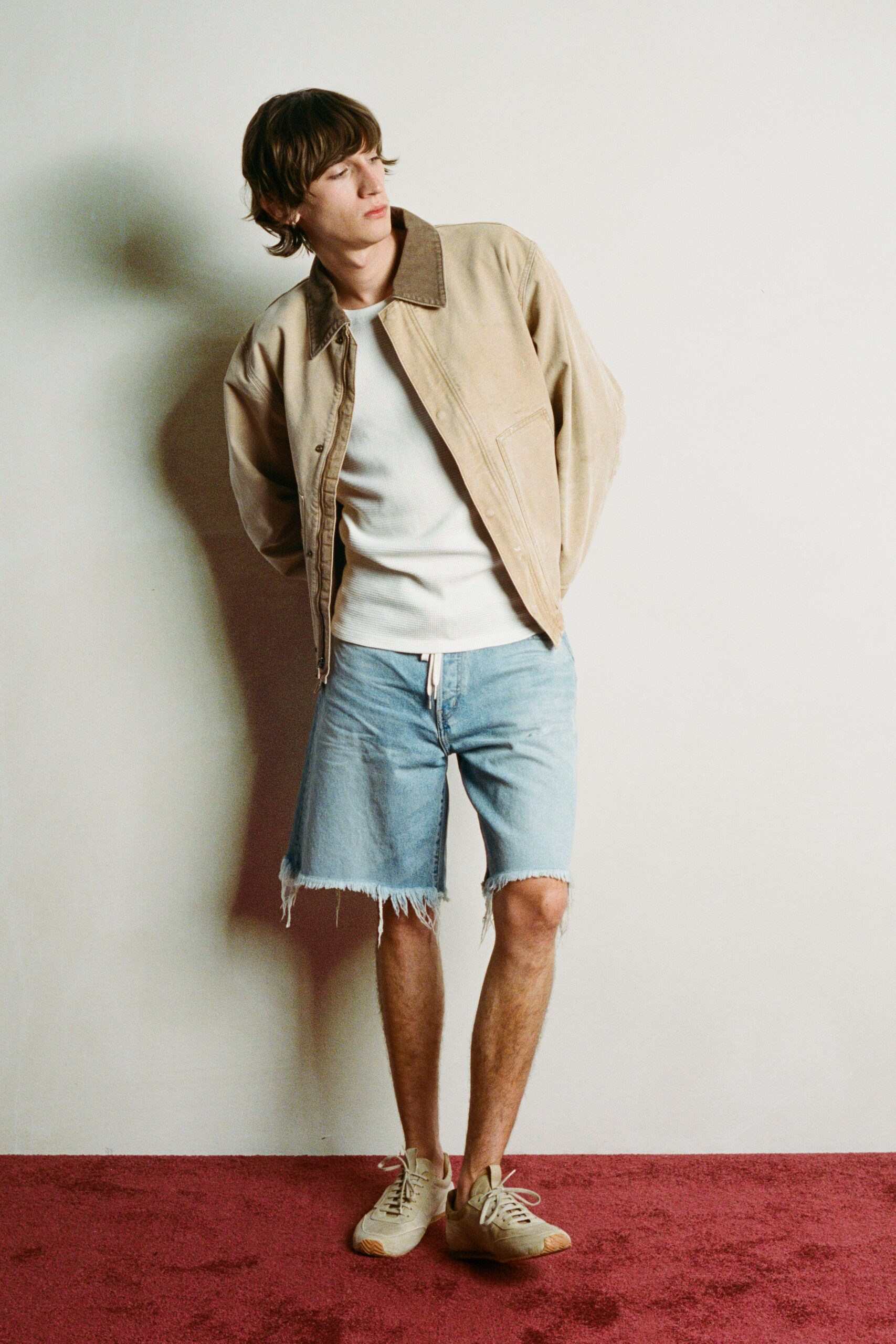 Stone canvas jacket with a darker corduroy collar and snap placket. White waffle-knit tee, close through the body. Pale denim cutoff shorts with long frayed threads at the hem. Cream low-top sneakers with gum-rubber toe caps. Walking back from the beach store with melted ice cream on your wrist.