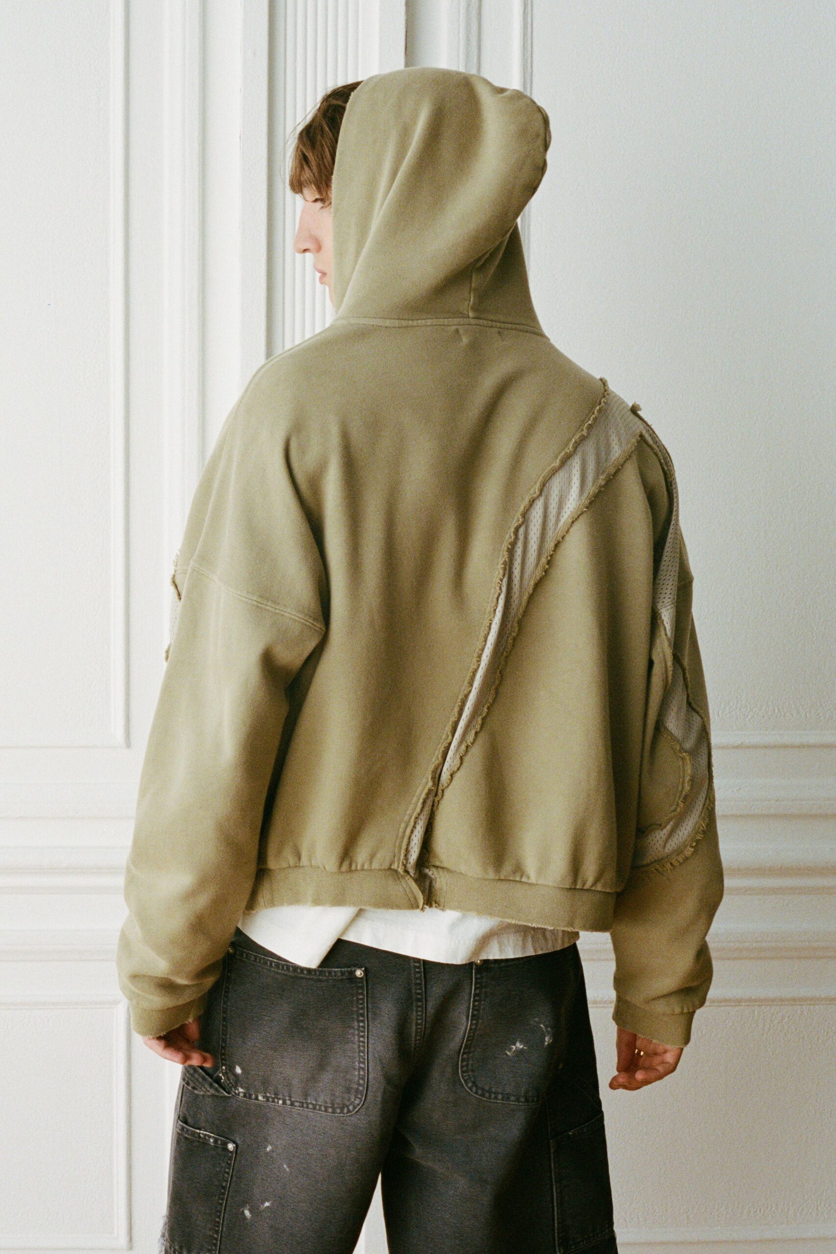 Khaki fleece hoodie with diagonal raw seams exposing cream mesh panels. White tee hem jutting out below the ribbed waistband. Black washed jeans with carpenter pockets and scattered white paint flecks. Thin gold band on one finger. Sunday apartment light, carrying rollers and a half-dry drop cloth.