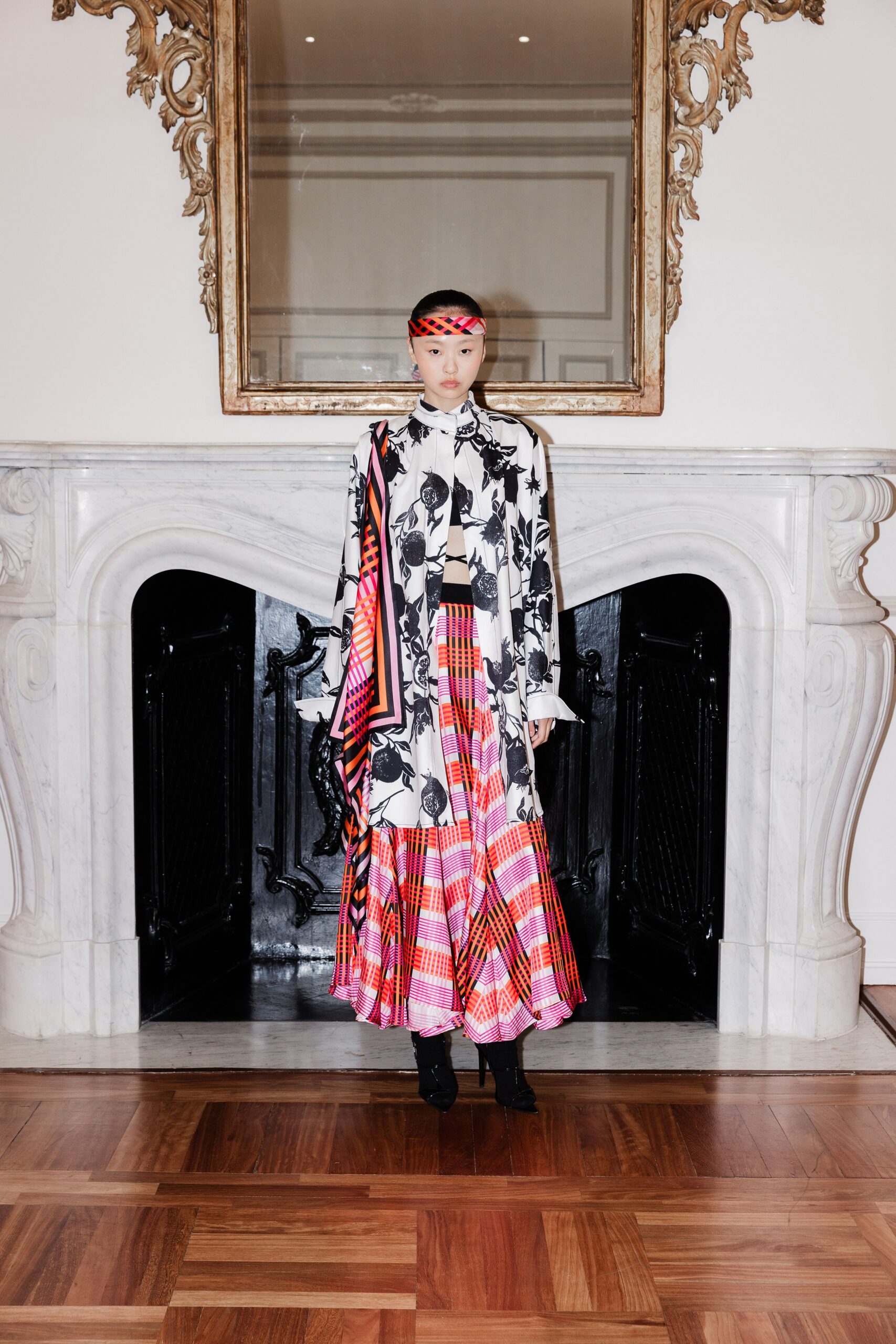 Neon orange and magenta lattice band worn flat across the forehead. White shirt dress printed with black pomegranates, one plaid panel falling from the shoulder. Floor-length plaid skirt in hot pink and orange, hem pooling wide. Black pointed ankle boots under the swaying hem. Crossing a marble hall before the house tour starts.