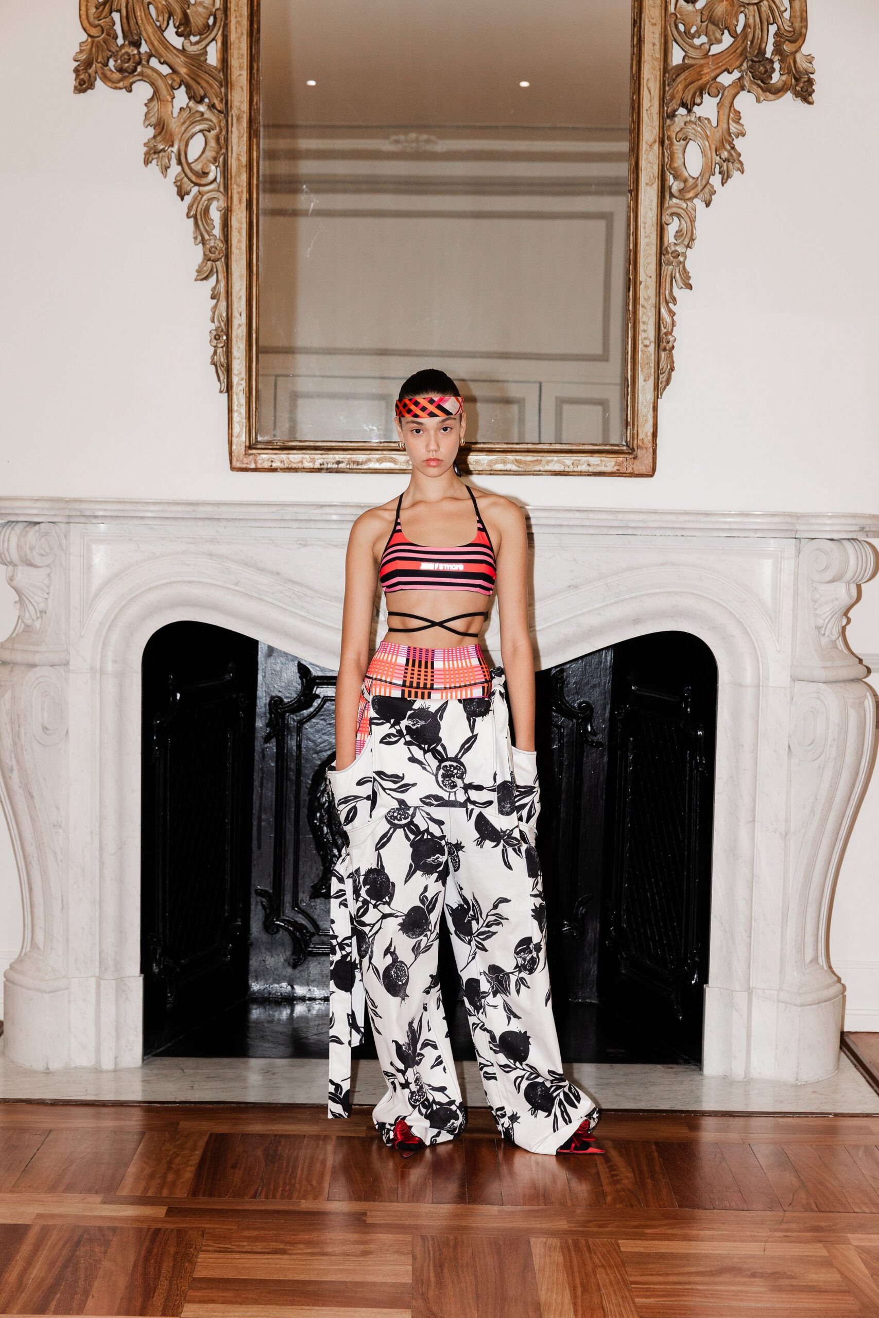 Neon plaid headband knotted flat across the forehead. Pink-and-black striped swim top with thin straps crossing the bare ribs. White wide-leg trousers printed with black pomegranates, pockets slouched open. Red satin heels peeking from the pooled hem. Crossing a marble hotel lobby still dressed for the pool.