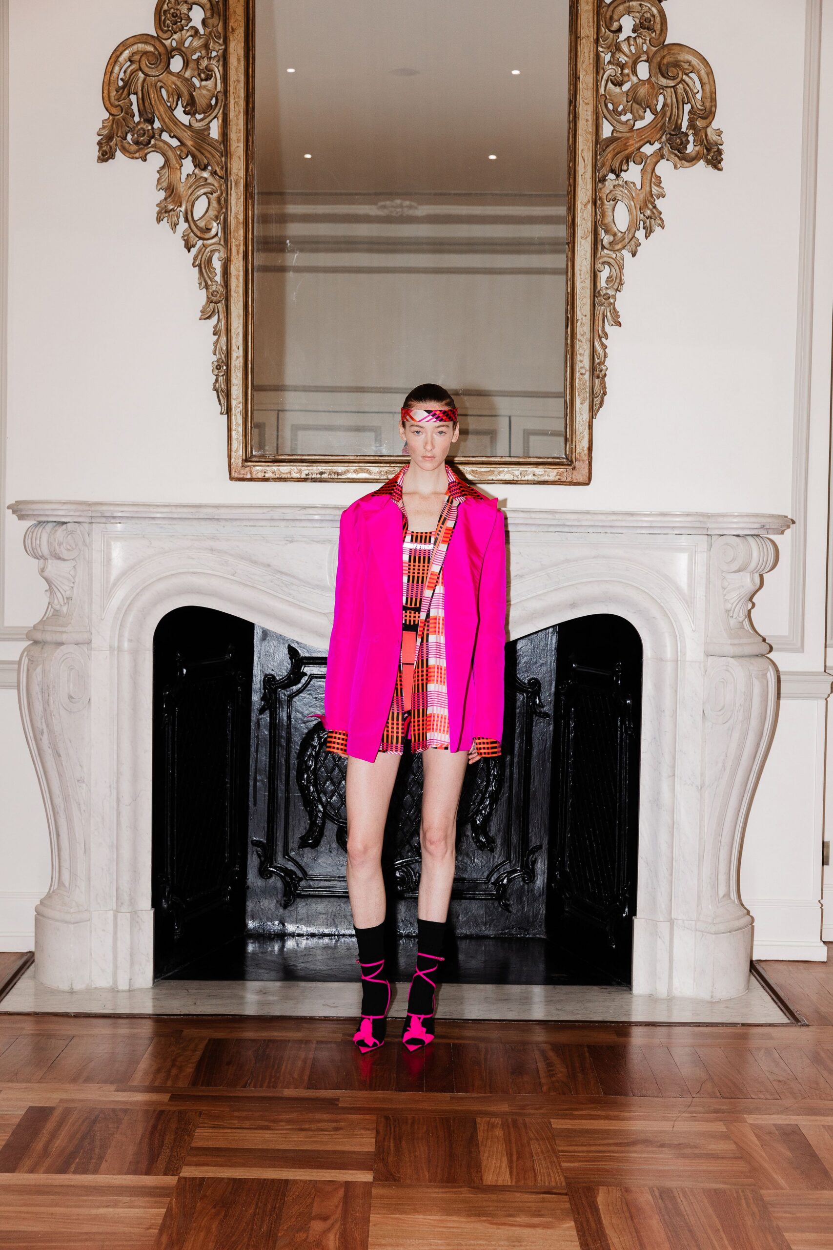 Narrow tartan headband worn flat across the forehead. Hot-pink blazer hanging open over a clashing plaid lapel. Orange-and-pink plaid playsuit with a square neckline and short hem. Black ankle socks. Fuchsia lace-up heels with cords spiraling over the socks. In a drawing room at noon, daring the carpet to clash back.