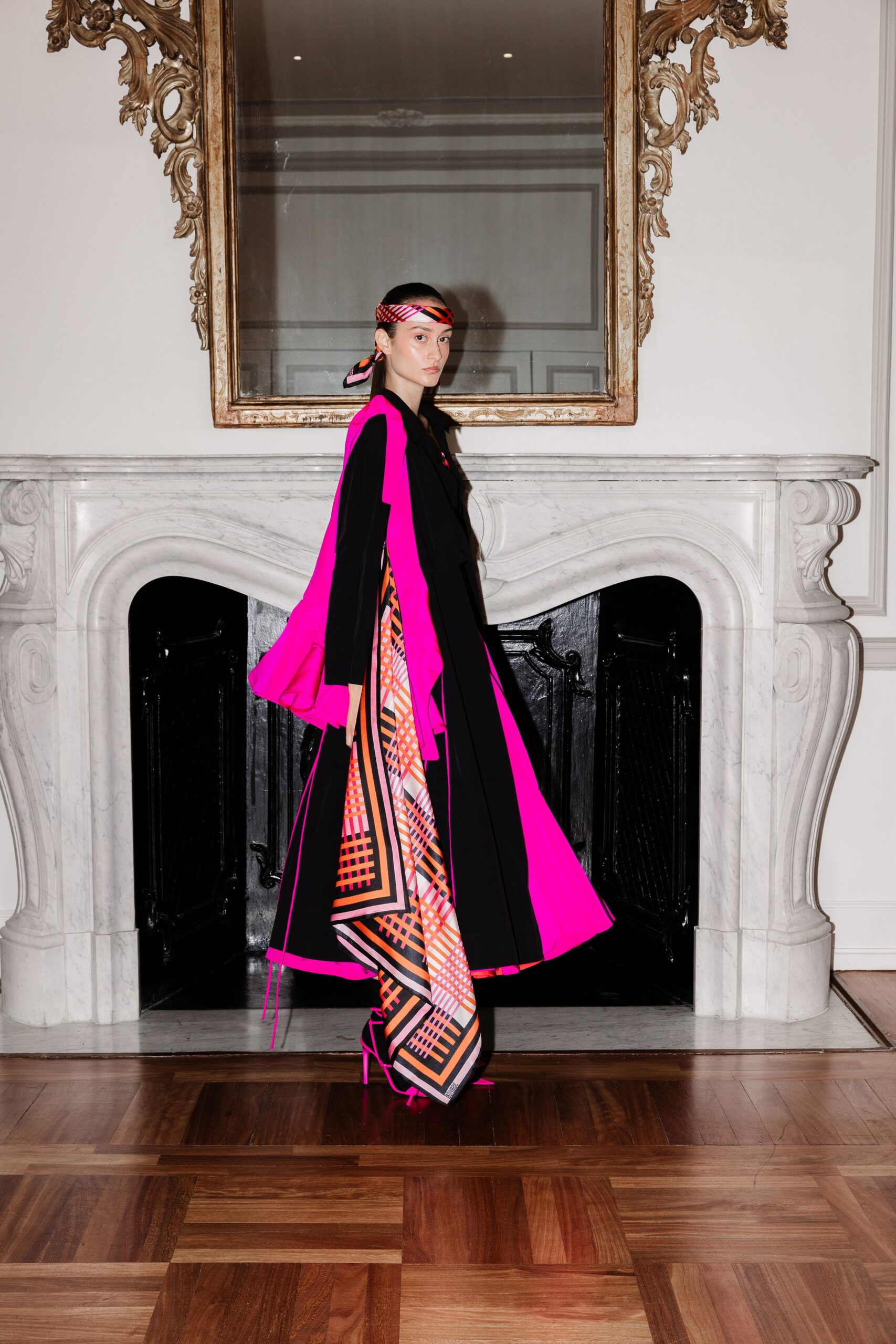 Narrow silk headscarf in coral and black checks, tails tied low. Black coat split open with blazing fuchsia lining and dangling side cords. Floor-length scarf-print panel dress, orange lattice framing the front slit. Hot-pink pumps with needle heels. Crossing a marble drawing room before anyone has finished setting the drinks.