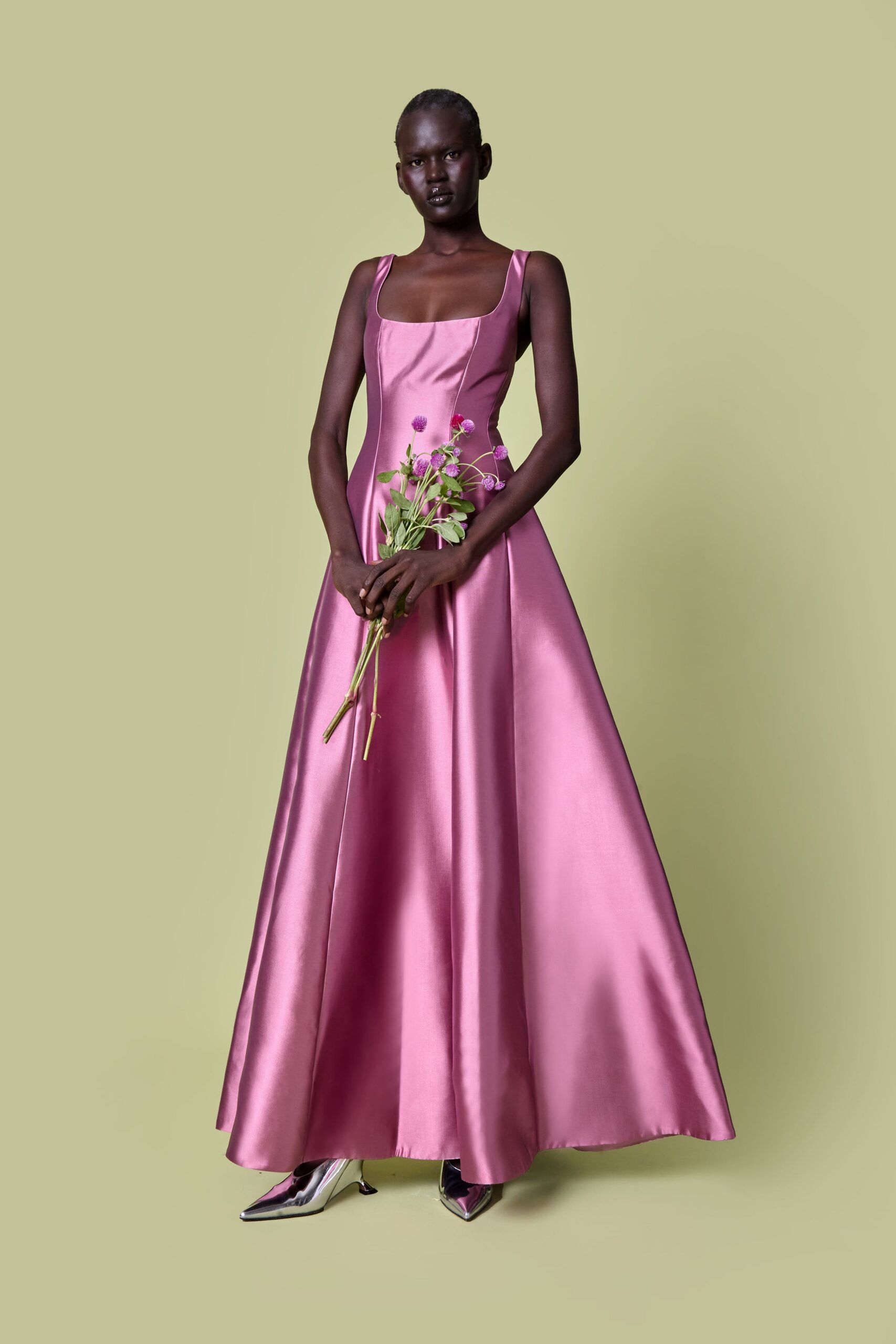 Rose satin ballgown with a square neckline and long sculpted panels. Mirror-silver pointed pumps under the hem, one toe catching light. Holding flowers after the city-hall wedding, before anyone finds the restaurant.