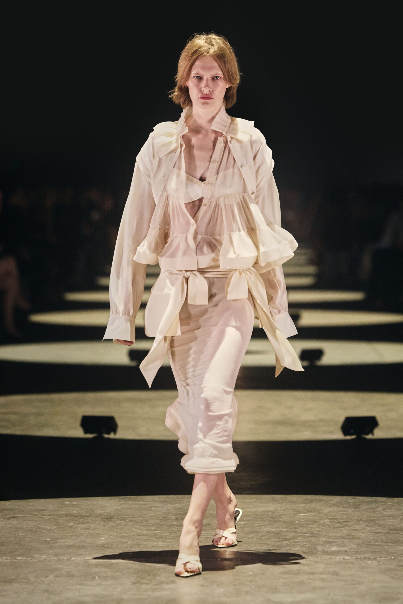 Sheer ivory blouse with tiered ruffles and a wide self-tie waist. Soft bandeau bralette peeking through the open placket. Semi-sheer ivory trousers, ruched in rippling vertical lines to the ankle. White crossover sandals with a slim kitten heel. Walking across a hotel atrium before breakfast service has started.