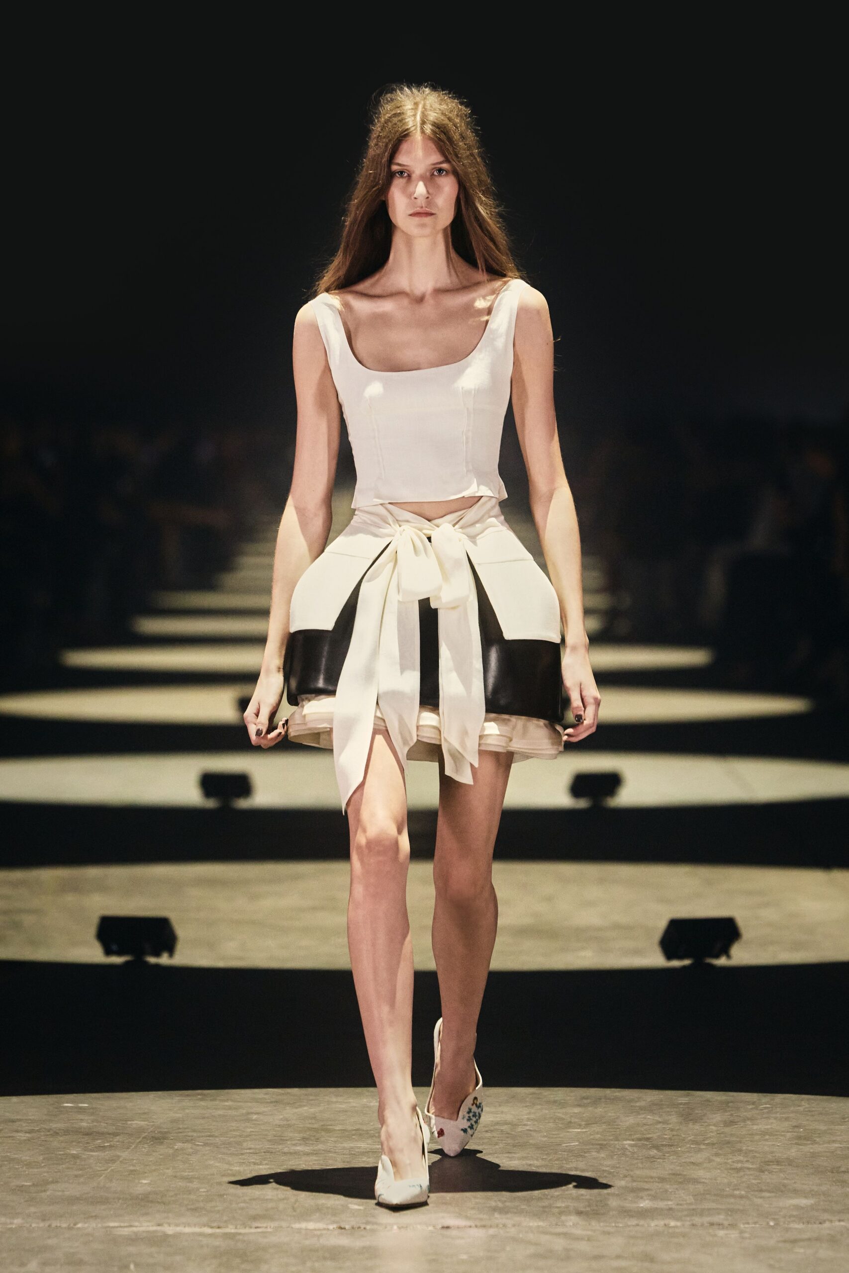 Ivory fitted top with a square scoop neckline and seamed cups. Ivory mini skirt tied in front, black bands curving around padded hips. Pointed white pumps with tiny floral embroidery at the vamp. For crossing a hotel atrium when the wedding brunch has already started.