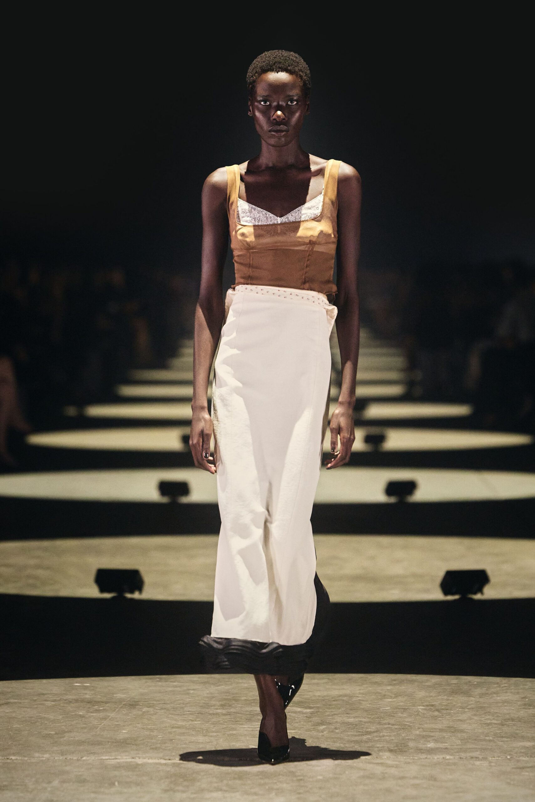 Ochre satin camisole with a square neckline framing white lace. Ivory column skirt, low waist picked out with tiny eyelets. Black patent pumps with a sharp pointed toe. Walking into a hotel restaurant before breakfast service has started.