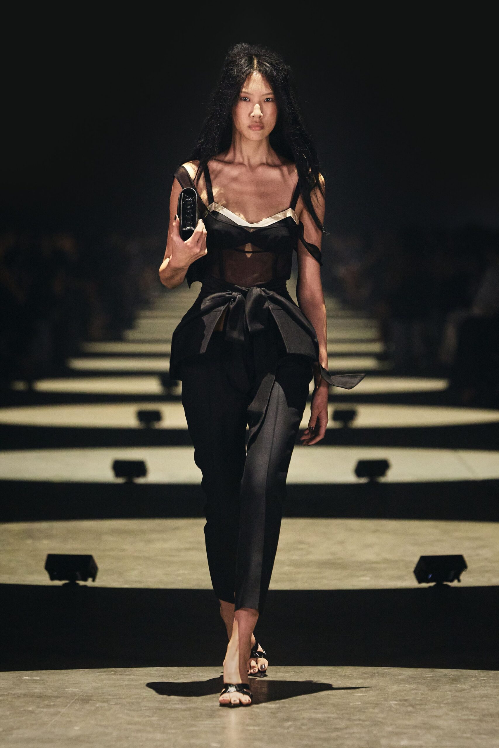 Sheer black camisole top with pale satin binding and one fallen strap. High-waisted black trousers, wrapped with a broad self-tie peplum bow. Glossy black box clutch gripped upright in one hand. Black patent thong sandals. Whatever you wear home when the hotel bar bill gets disputed at dawn.