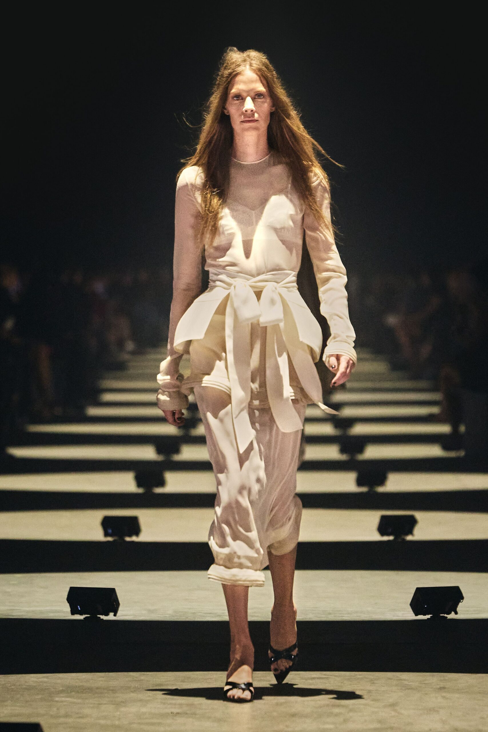 Sheer nude long-sleeved dress with opaque ivory appliqué and a plunging inset. Wide self-fabric bow cinched hard, tails hanging past the hips. Black patent slingback heels with a sharply pointed toe. Crossing a museum atrium after midnight, still lit from below.