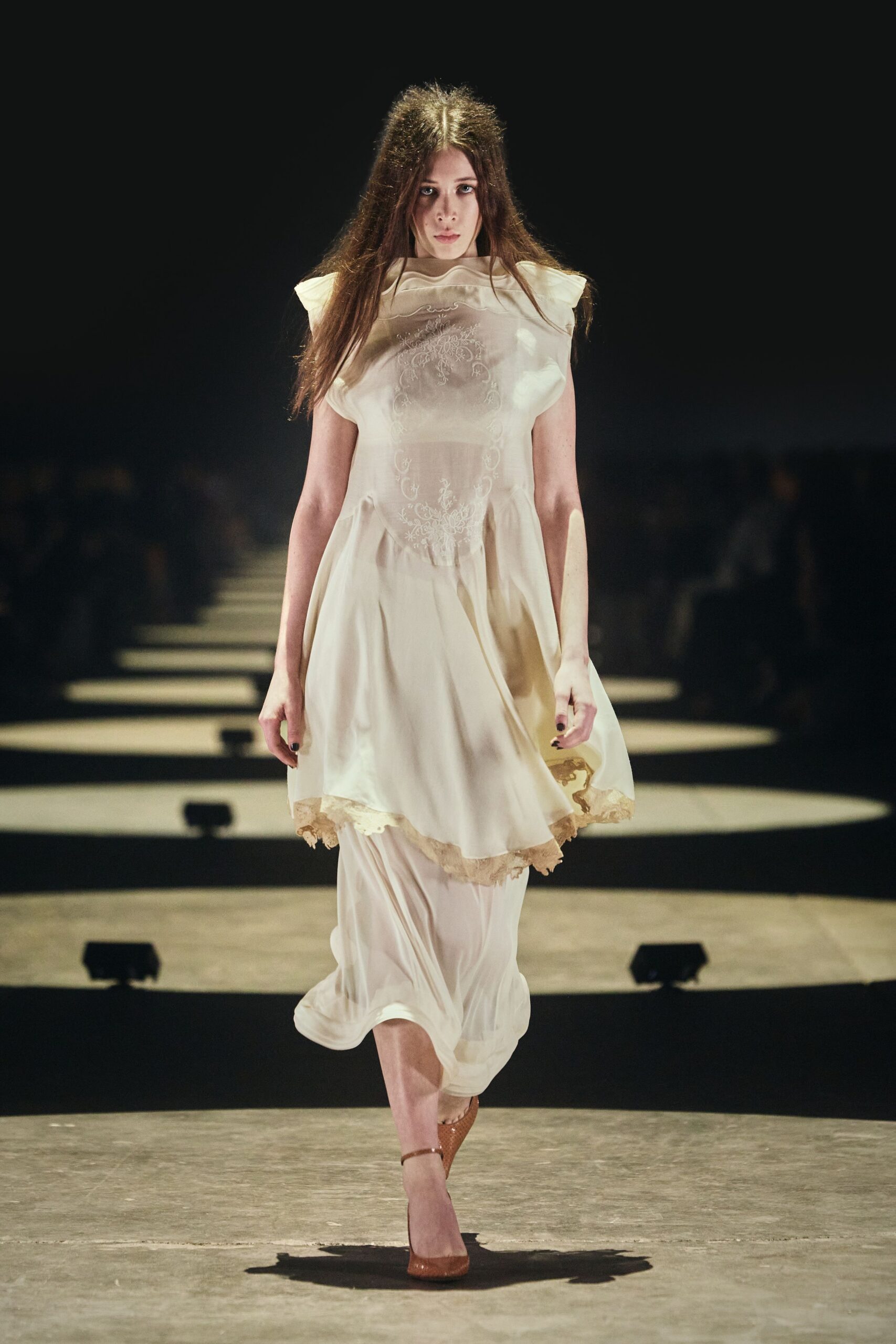 Ivory chiffon dress with a sheer embroidered bib and tucked waist. Longer underlayer pooling below, its lace hem peeking at the side. Tan ankle-strap pumps in pebbled leather. Crossing a candlelit hall after the wedding guests have gone upstairs.