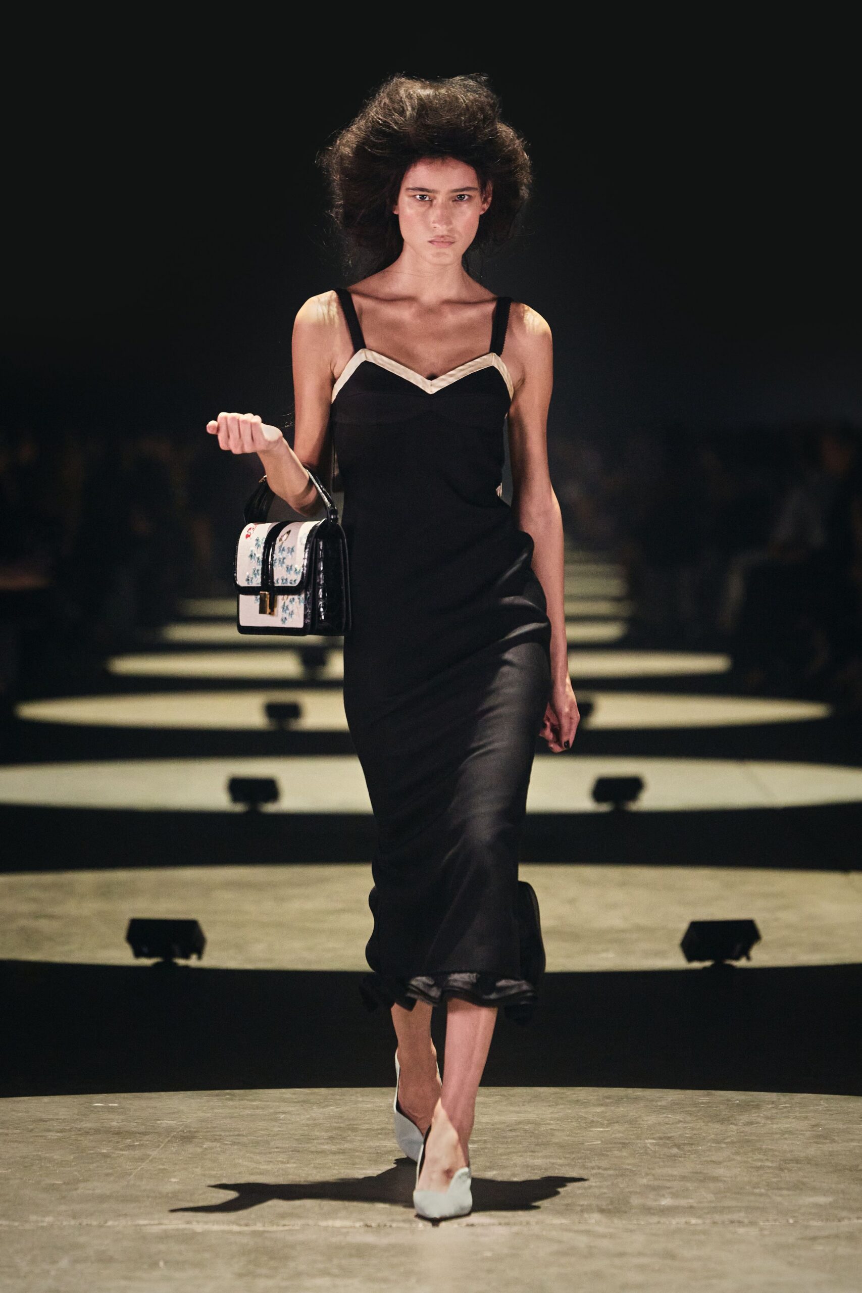 Black bias-cut slip dress with ivory satin chevrons at the neckline. Boxy black patent bag with porcelain-floral panels and a square clasp. Powder-blue pointed pumps, the left toe lightly creased. After the wedding dinner, carrying the cake knife back through the hotel corridor.