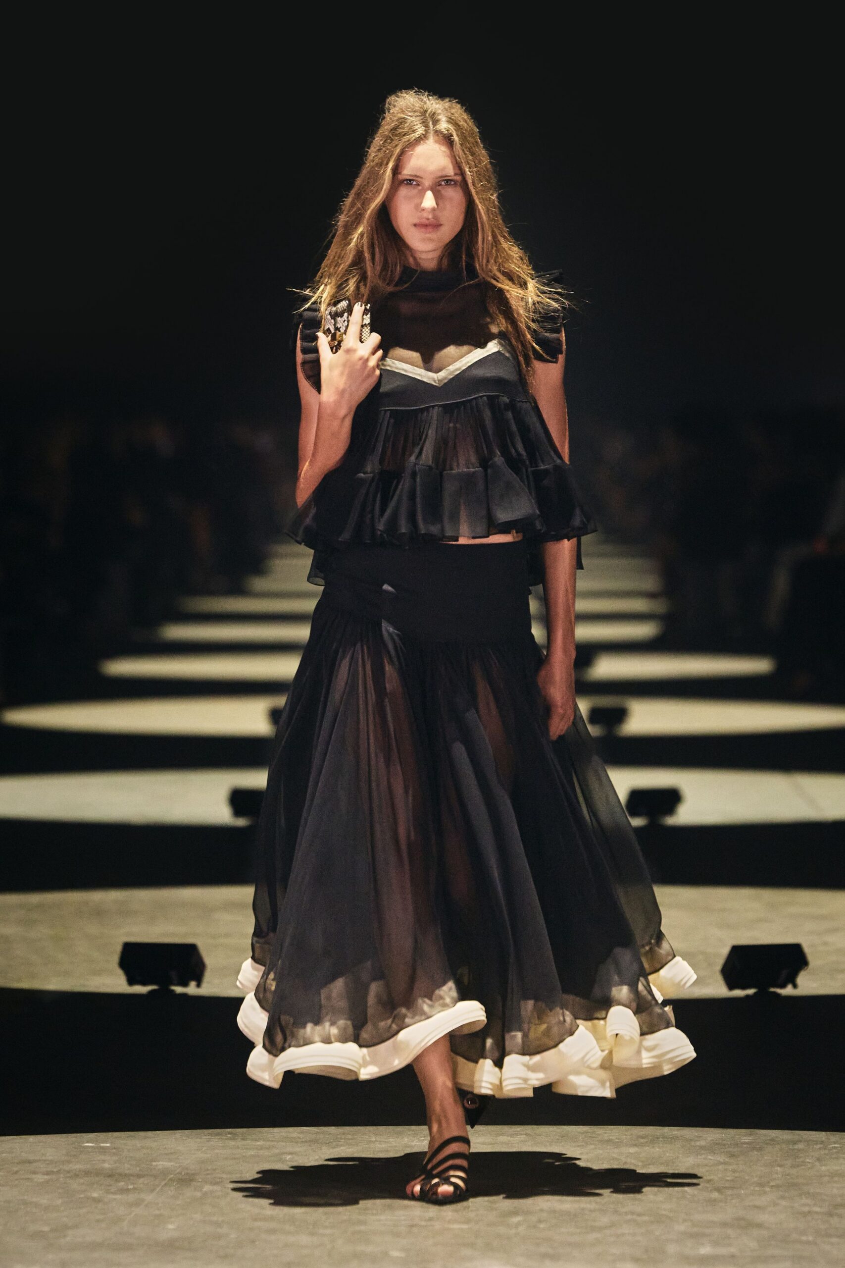 Black sheer-mesh yoke over a sweetheart bodice, tiered chiffon flounces at the waist. Full black chiffon skirt, softly pleated with a thick ivory rolled hem. Small python clutch tucked under one arm. Black strappy sandals. Crossing a hotel ballroom after the wedding band has packed up.