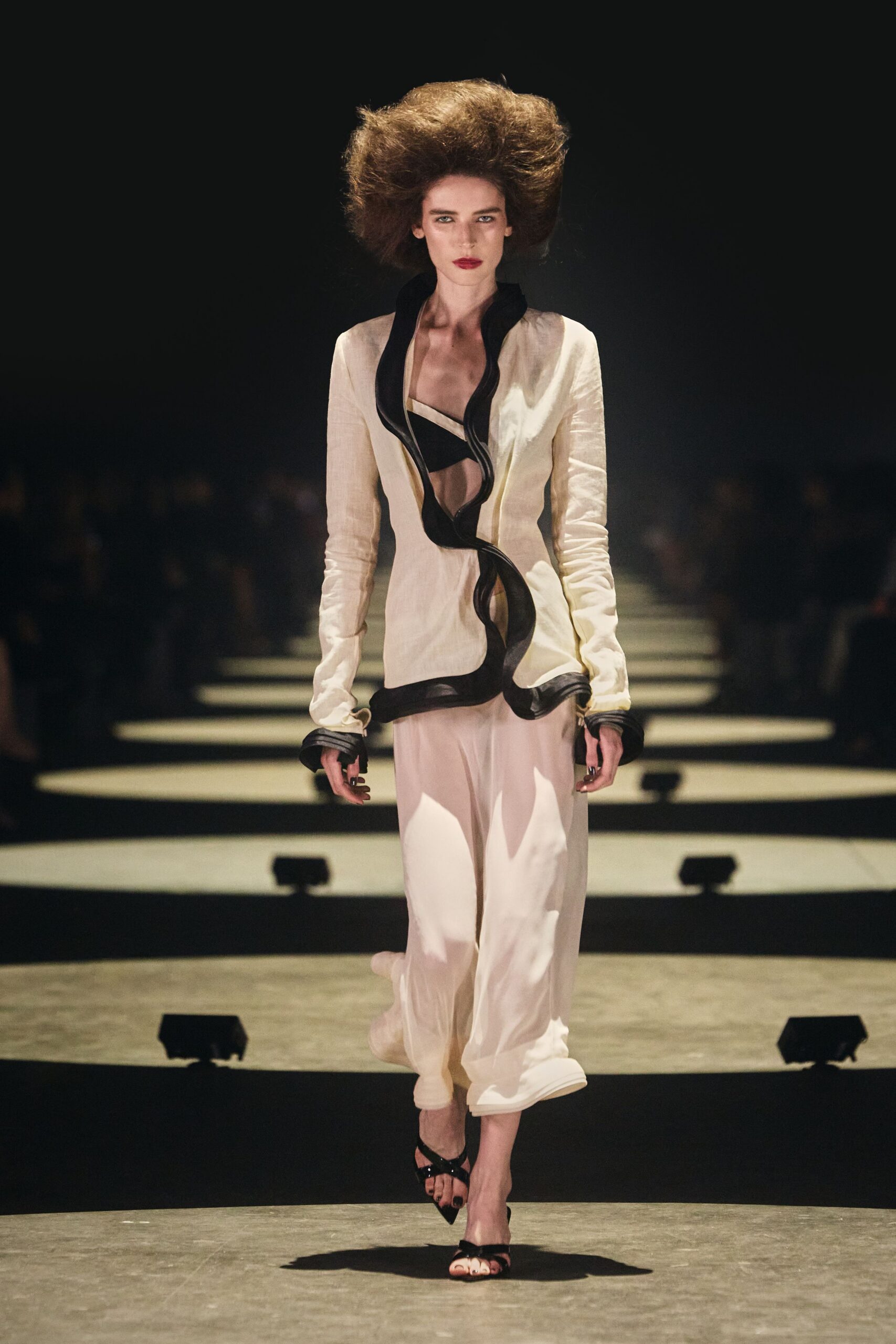 Ivory shantung jacket cut open to the navel, black ruffle snaking downward. Black bandeau bralette with a narrow pale binding at the top edge. Blush silk trousers falling loose and sheer over the ankles. Black patent toe-ring sandals. Walking hotel corridors after a wedding, carrying your shoes by dawn.