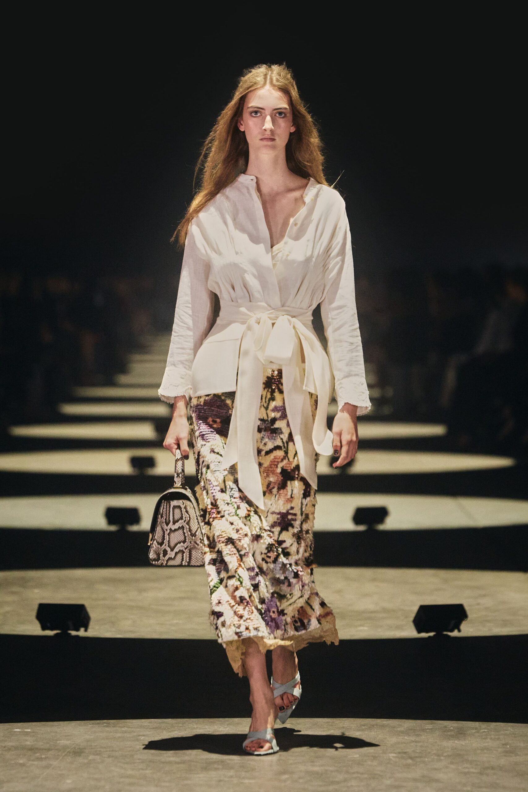 Ivory blouse in crushed silk, gathered deep and tied in a wide sash. Cream camisole peeking at the open placket. Bias floral brocade skirt, frayed lace hem brushing the ankles. Small python top-handle bag. Pale crossed-strap heels. Leaving a vineyard lunch after the tablecloths have gone wine-stained.