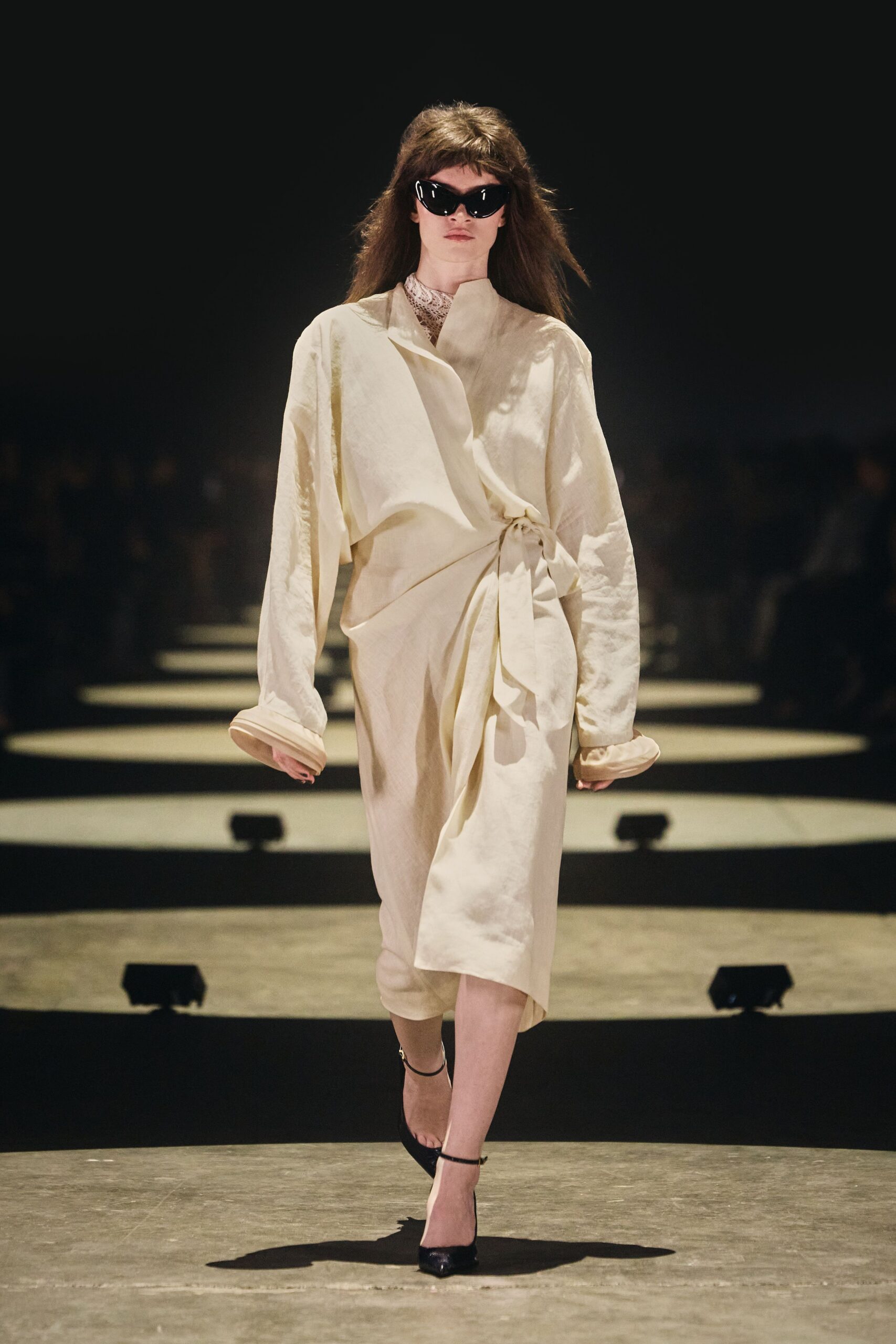 Glossy black cat-eye frames with thick temples. Cream satin wrap dress, side knot pulling the skirt into soft folds. Fine openwork lace peeking high at the throat. Black ankle-strap pumps with pointed toes. Leaving a courthouse wedding before anyone suggests champagne.
