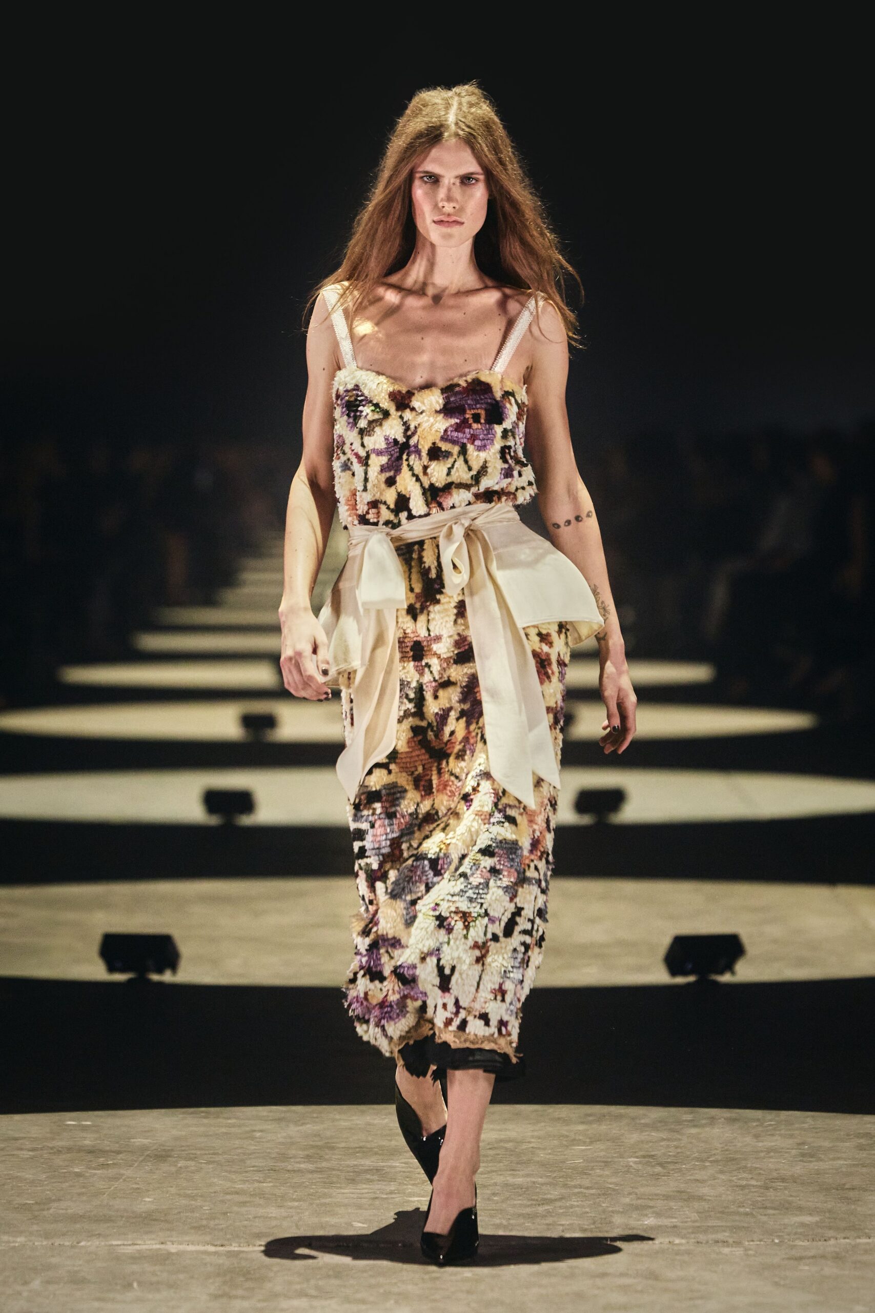 Ivory floral chenille column dress with narrow straps and a fuzzy nap. Sheer cream organza tied low, one loop collapsing over the hip. Black patent pumps. Walking through a hotel ballroom after the chairs have been stacked.