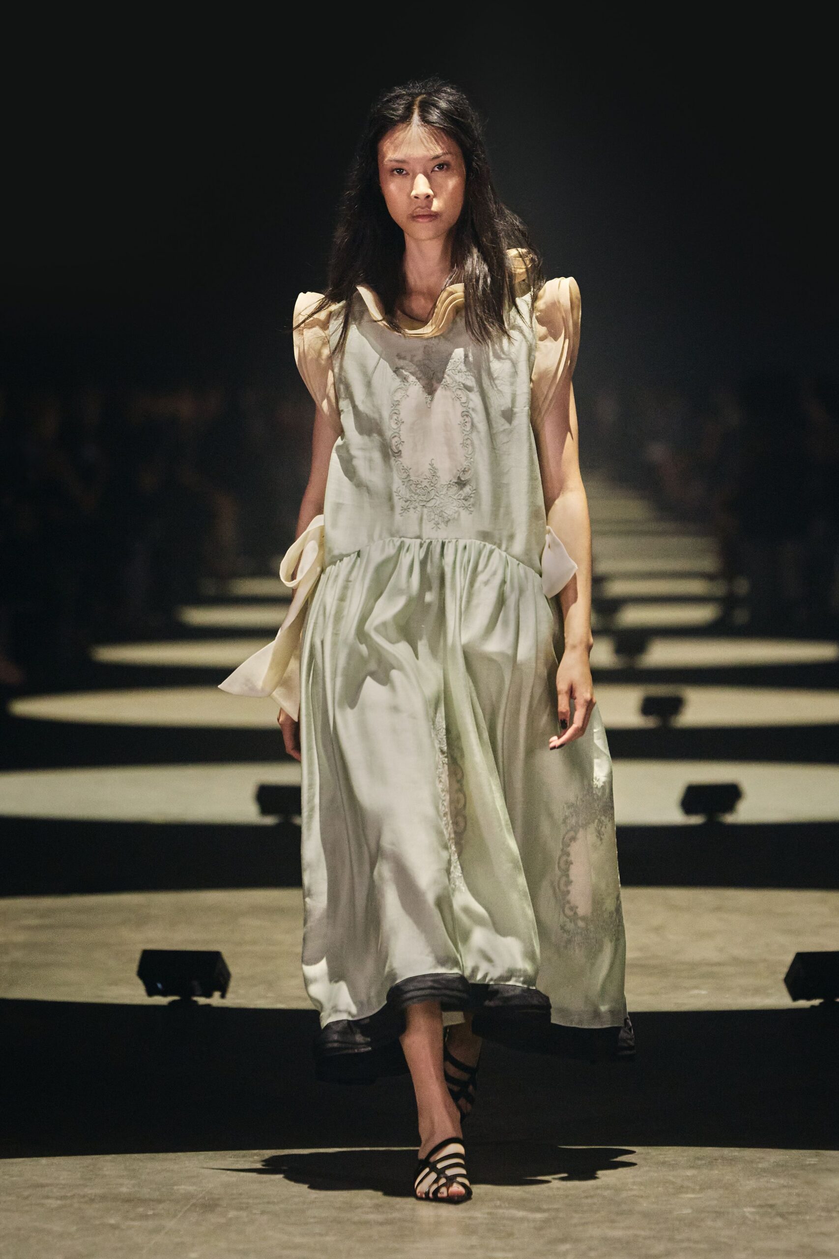 Pale satin shift dress with a dropped gathered waist and ghosted embroidery. Buttercream organza flounces standing off the shoulders in soft curls. Black strappy sandals, narrow bands crossing the toes. Walking the long hotel corridor after a wedding, carrying your shoes by midnight.