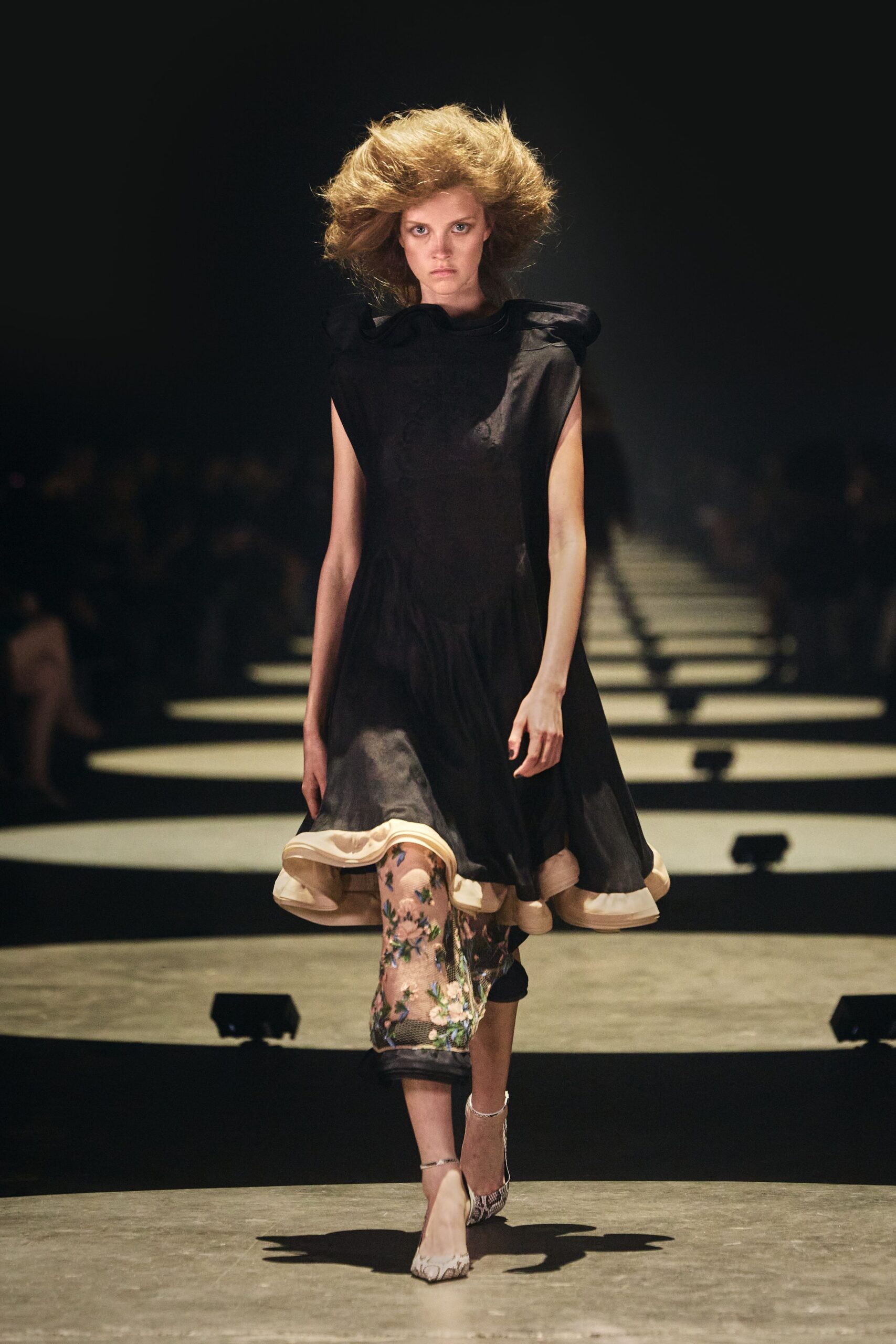 Black satin dress with puffed shoulders and a cream-faced hem kick. One nude mesh leg embroidered with blue and cream flowers. Pointed snake-print heels with slim silver ankle straps. Crossing a hotel ballroom after midnight, carrying the corsage from dinner.