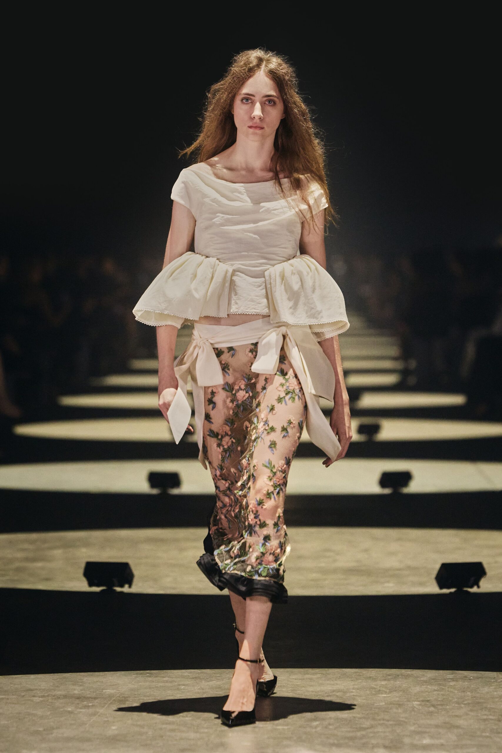 Ivory top with a draped bateau neck and pannier-like peplum sleeves. Wide cream sash tied low, one bow hanging past the hip. Blush mesh pencil skirt densely embroidered with climbing flowers. Black patent pumps with a fine ankle strap. For reading a note in the conservatory after everyone else has gone.