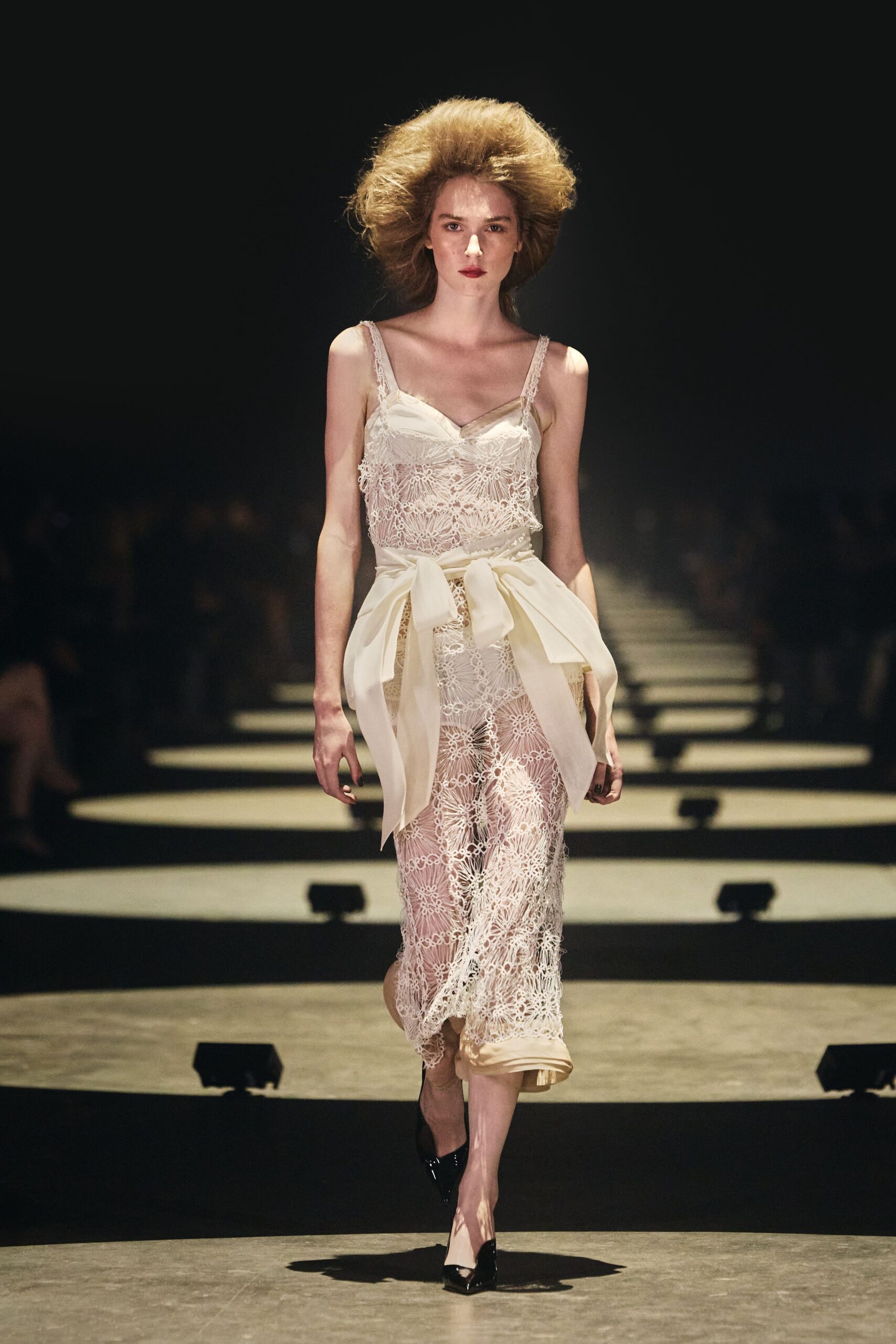 Ivory floral lace slip dress, satin cups and straps peeking through. Wide ivory sash tied low, the bow tails hanging past the hips. Black patent pumps with narrow pointed toes. After the courthouse wedding, waiting for takeaway on a too-bright corner.