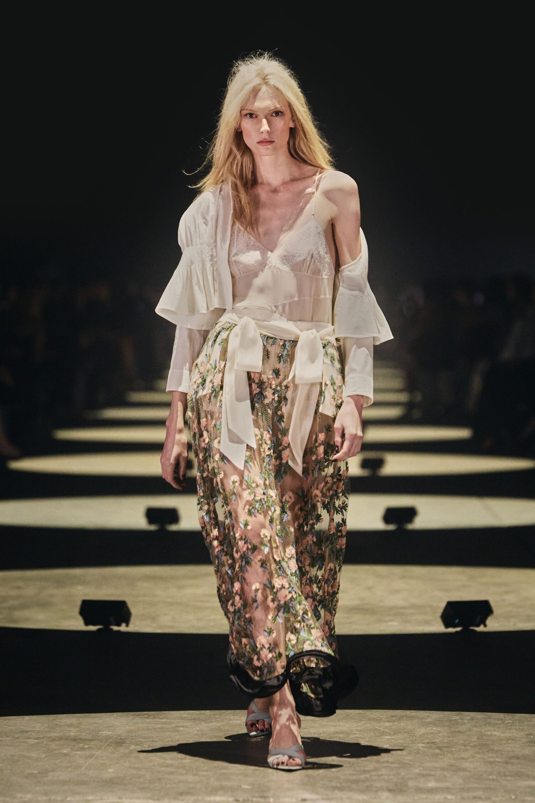 Ivory silk robe top slipping off one shoulder, tiered flute cuffs. Blush slip dress with lace cups and a satin bow knotted low. Sheer mesh skirt densely embroidered with pink blossoms over black hem. Pale blue toe-band sandals. Crossing a hotel courtyard at dawn with last night's flowers still pinned on.