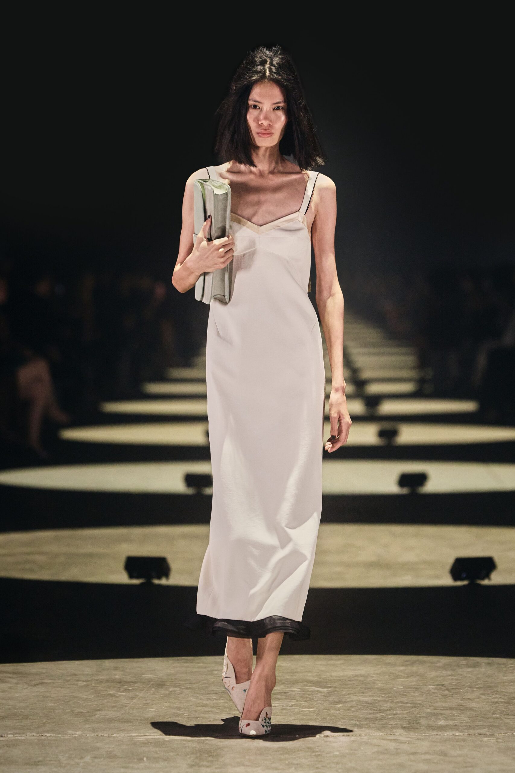 Ivory satin slip dress with a shallow V and black picot straps. Soft rectangular clutch tucked flat under one arm. White pointed pumps printed with tiny scattered sprigs. Walking from a registry office with the flowers left behind.